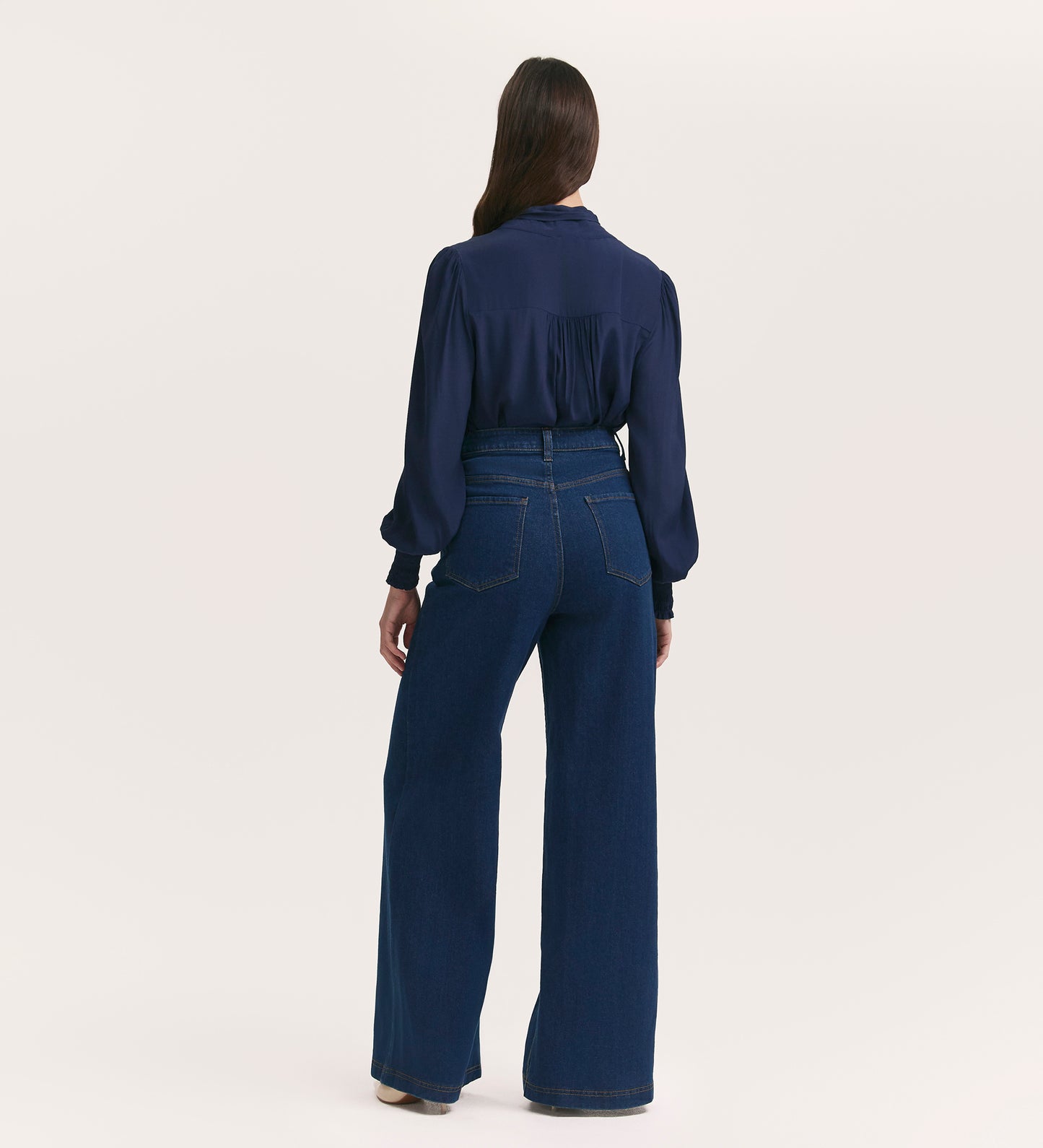 A woman with long brown hair faces away, wearing a navy long-sleeve blouse and Monica Mid Wash Wide Leg Jeans against a plain light background.