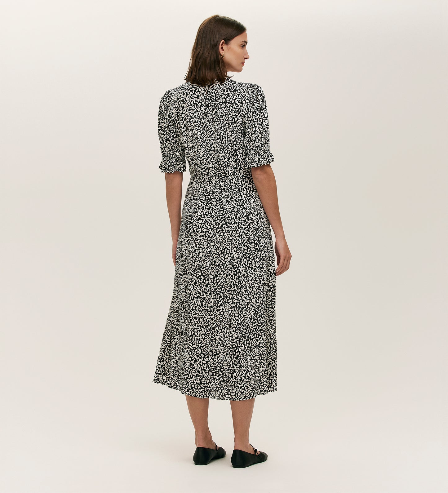 A woman with straight brown hair faces away, wearing the Mela Black Leopard Crepe Midi Tea Dress with short sleeves and black flats, set against a plain light background.