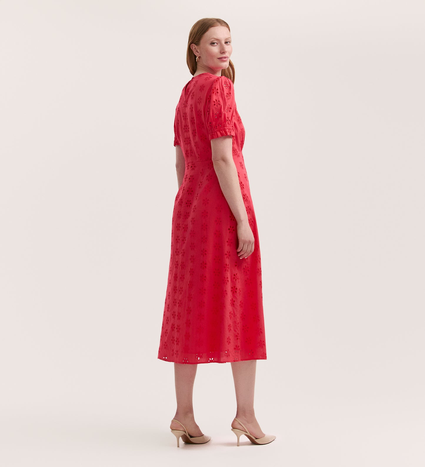 A woman with straight, light brown hair stands sideways in the Mela Coral Cotton Midi Tea Dress, featuring subtle embroidery, paired with beige low-heeled shoes. She faces the camera against a plain background.
