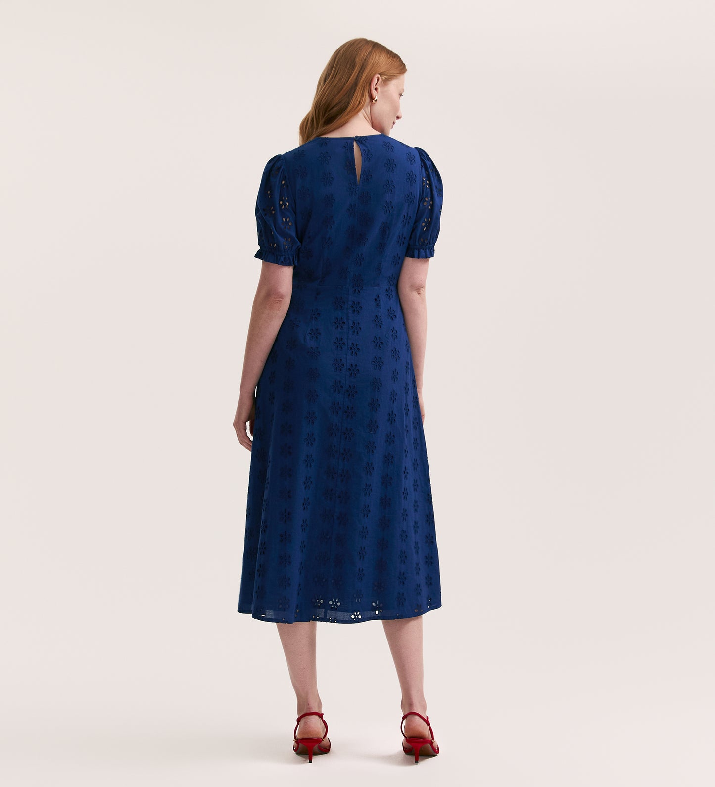 A woman with long red hair faces away against a plain light background, wearing the Mela Indigo Cotton Midi Tea Dress with embroidered eyelet details and red heeled sandals.