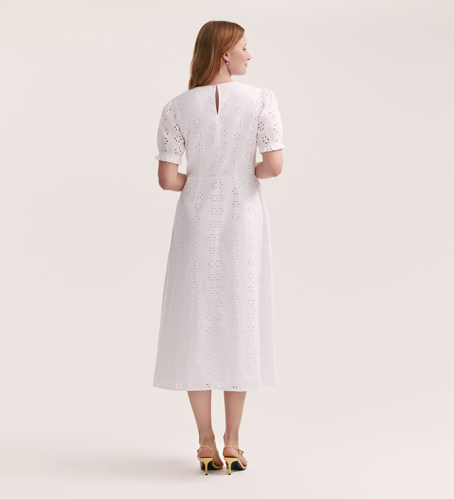 A woman faces away, wearing the Mela White Cotton Midi Tea Dress—an embroidered, empire-waist, calf-length cotton piece with a small back button detail—paired with yellow heeled sandals against a plain light backdrop.