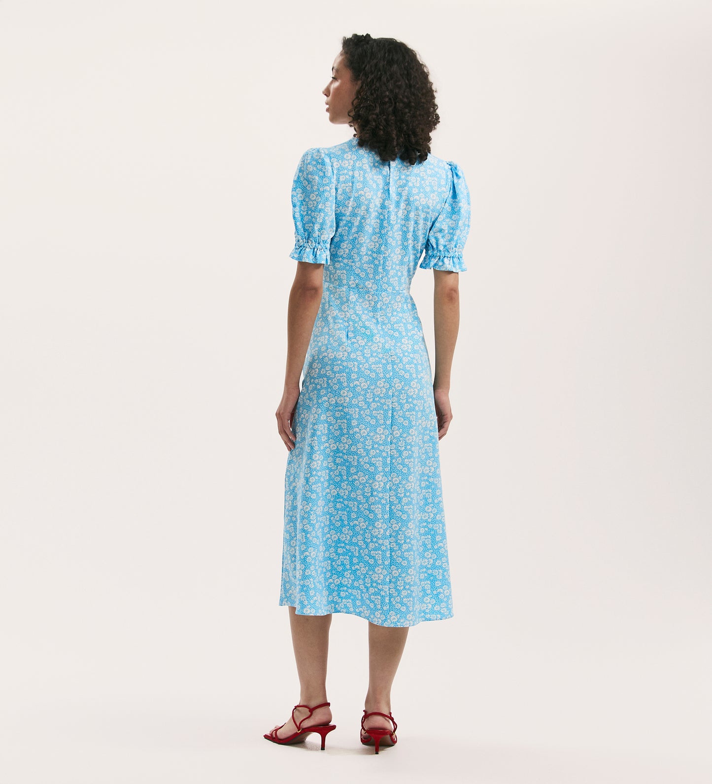 A woman faces away, modeling the Mela Blue Daisy Crepe Midi Tea Dress with an empire waist and short sleeves. Her curly hair and red heeled sandals stand out against a plain white background.