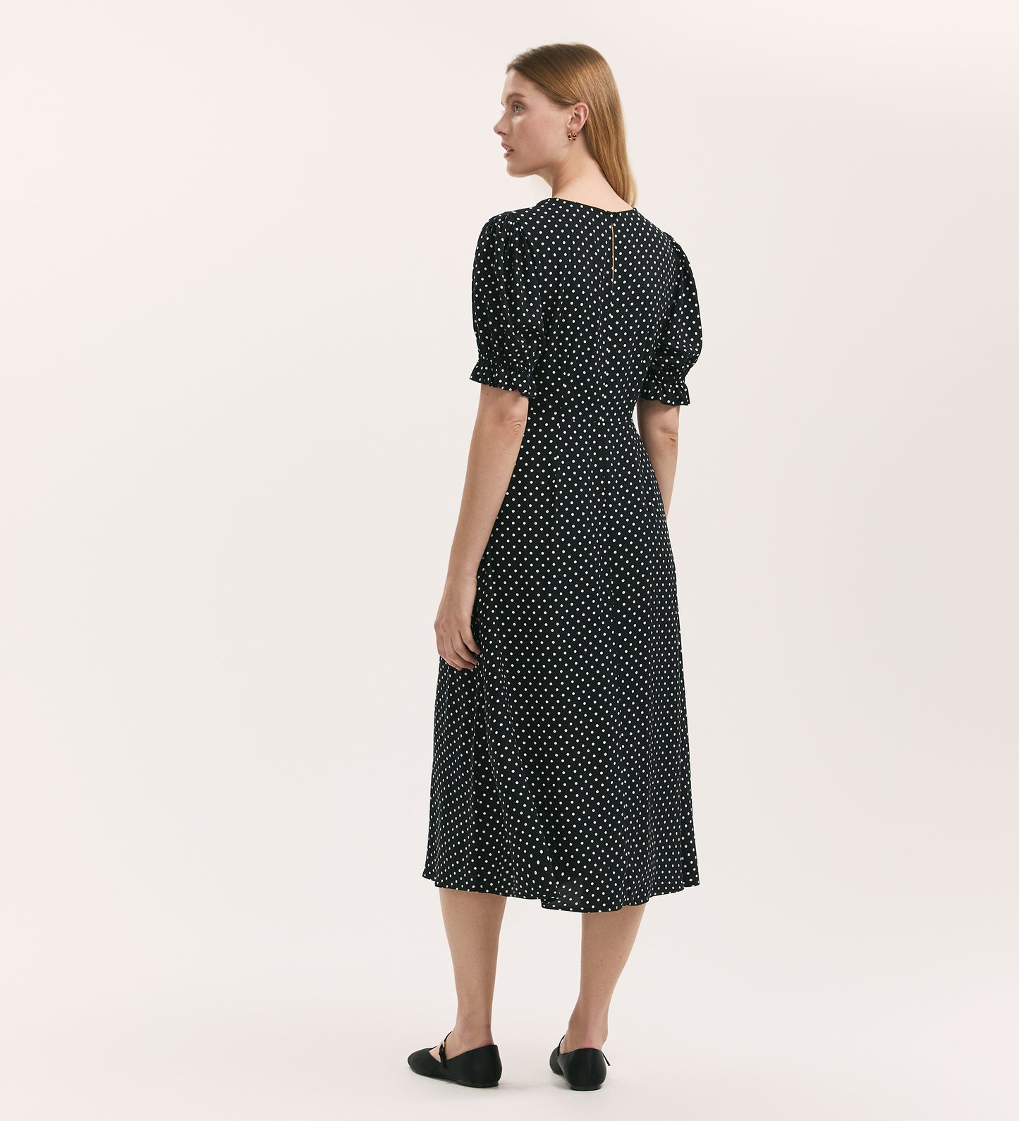 A woman faces away, wearing the Mela Black Polka Dot Midi Tea Dress with a round neckline and short puffed sleeves, teamed with black flats. The background is plain and light-colored.
