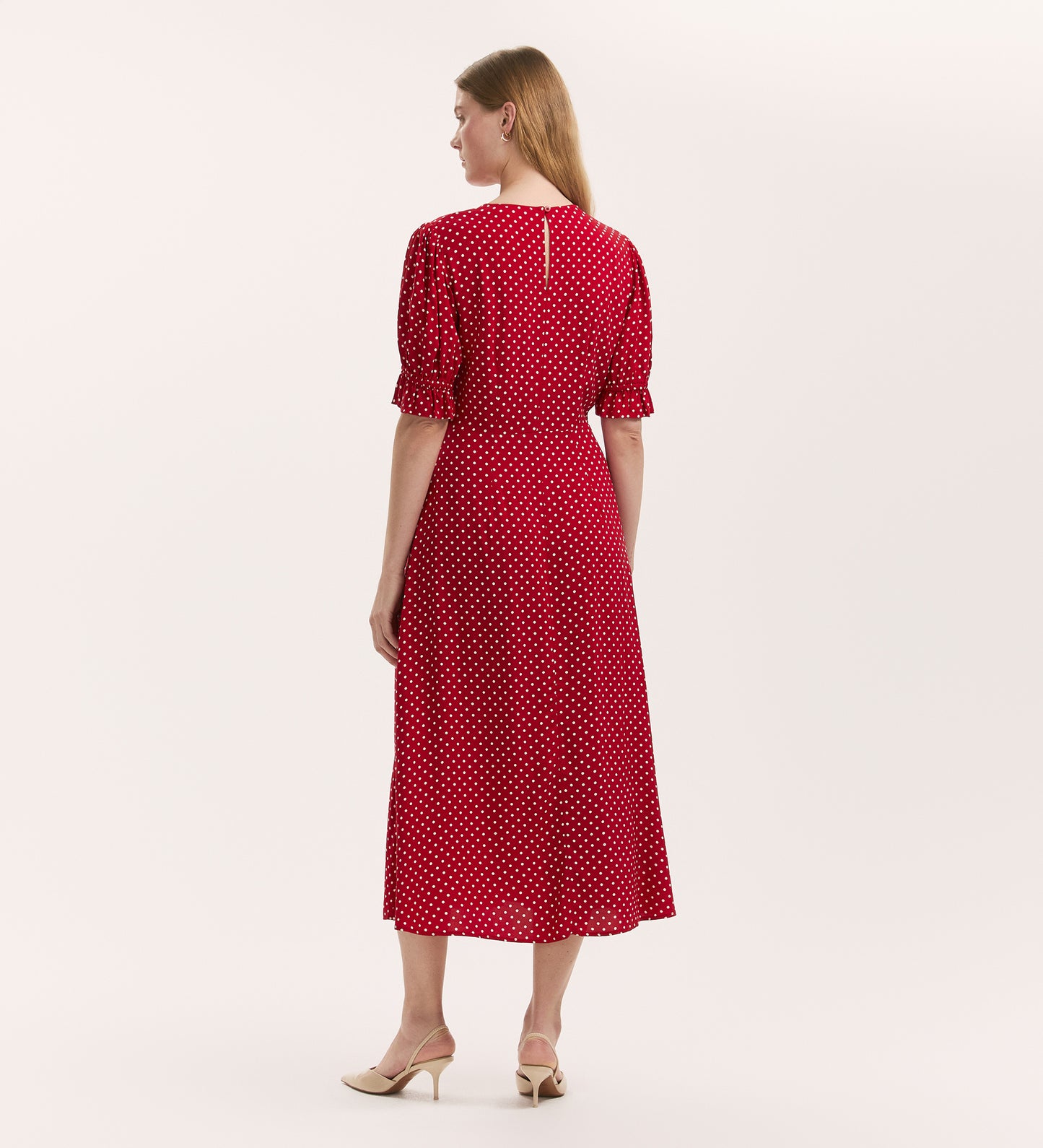 A woman, seen from behind, wears the Mela Red Polka Dot Midi Tea Dress—featuring white polka dots, short puffed sleeves, a round neckline with rear keyhole fastening—and beige low-heeled shoes against a plain white background.