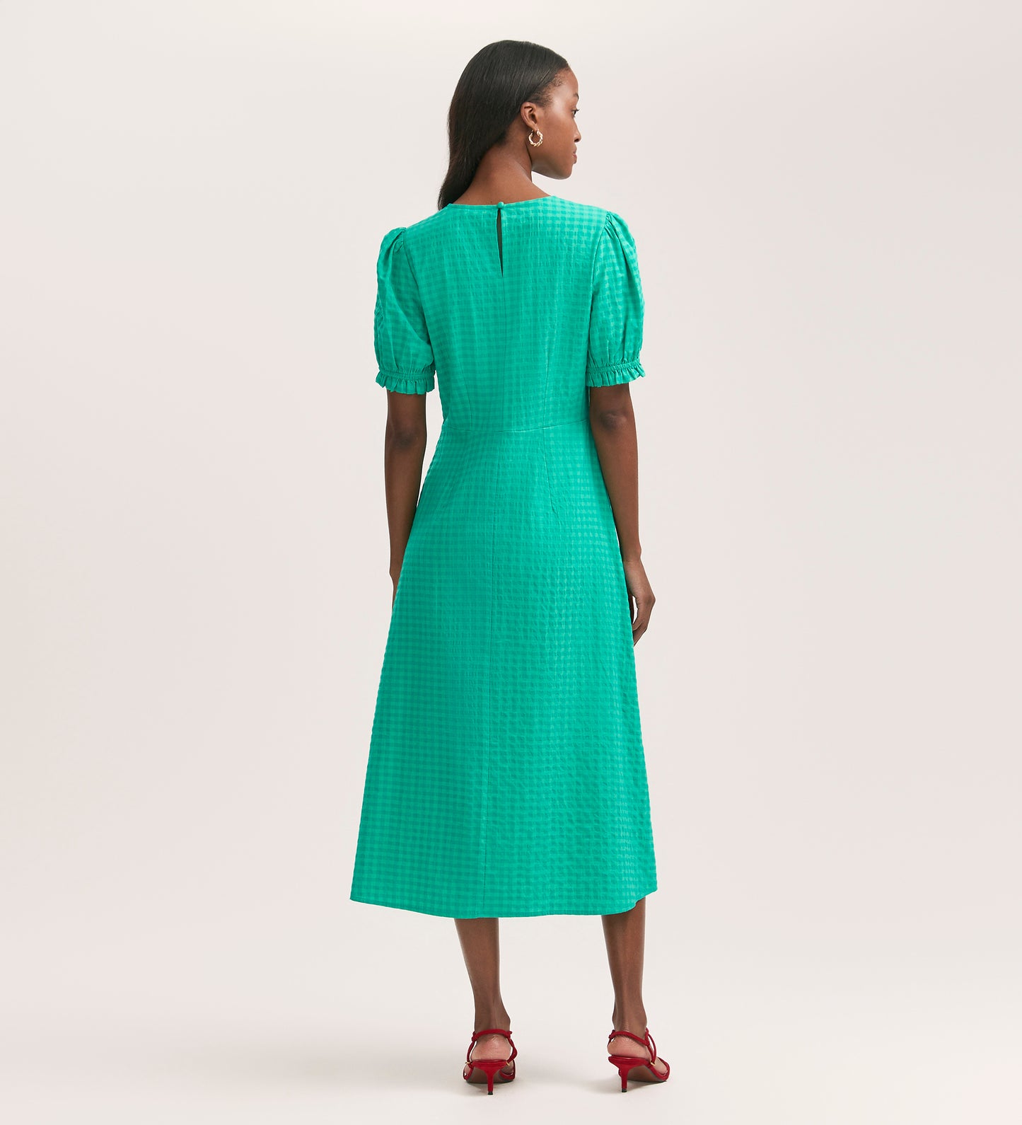 A woman with long dark hair faces away, wearing the Mela Green Gingham Seersucker Midi Dress featuring short puff sleeves and a subtle checkered pattern. She pairs it with red heeled sandals against a plain, light background.