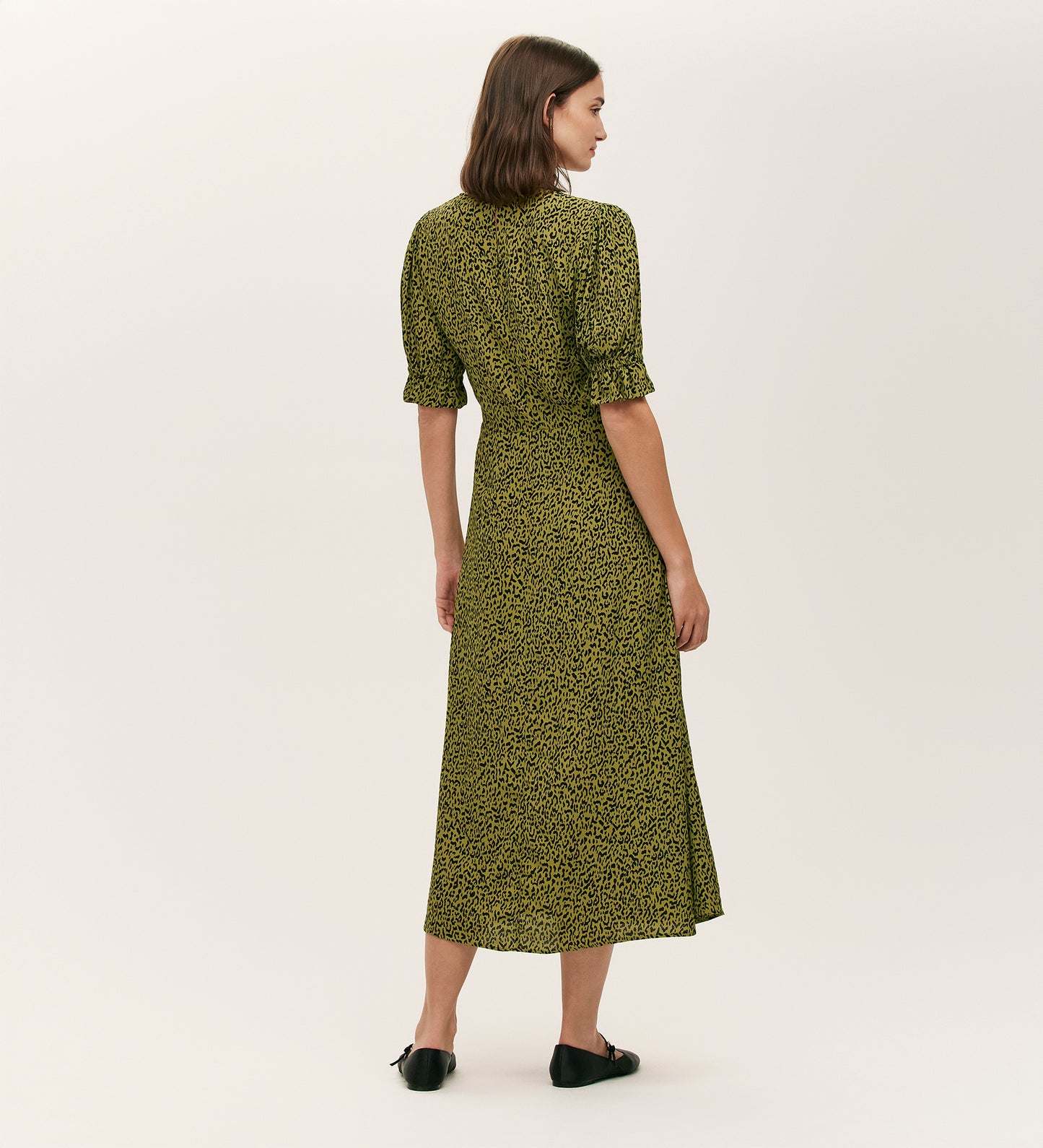 A woman with straight brown hair stands facing away, wearing the Mela Green Leopard Crepe Midi Tea Dress, short sleeves, paired with black flat shoes against a plain light background.