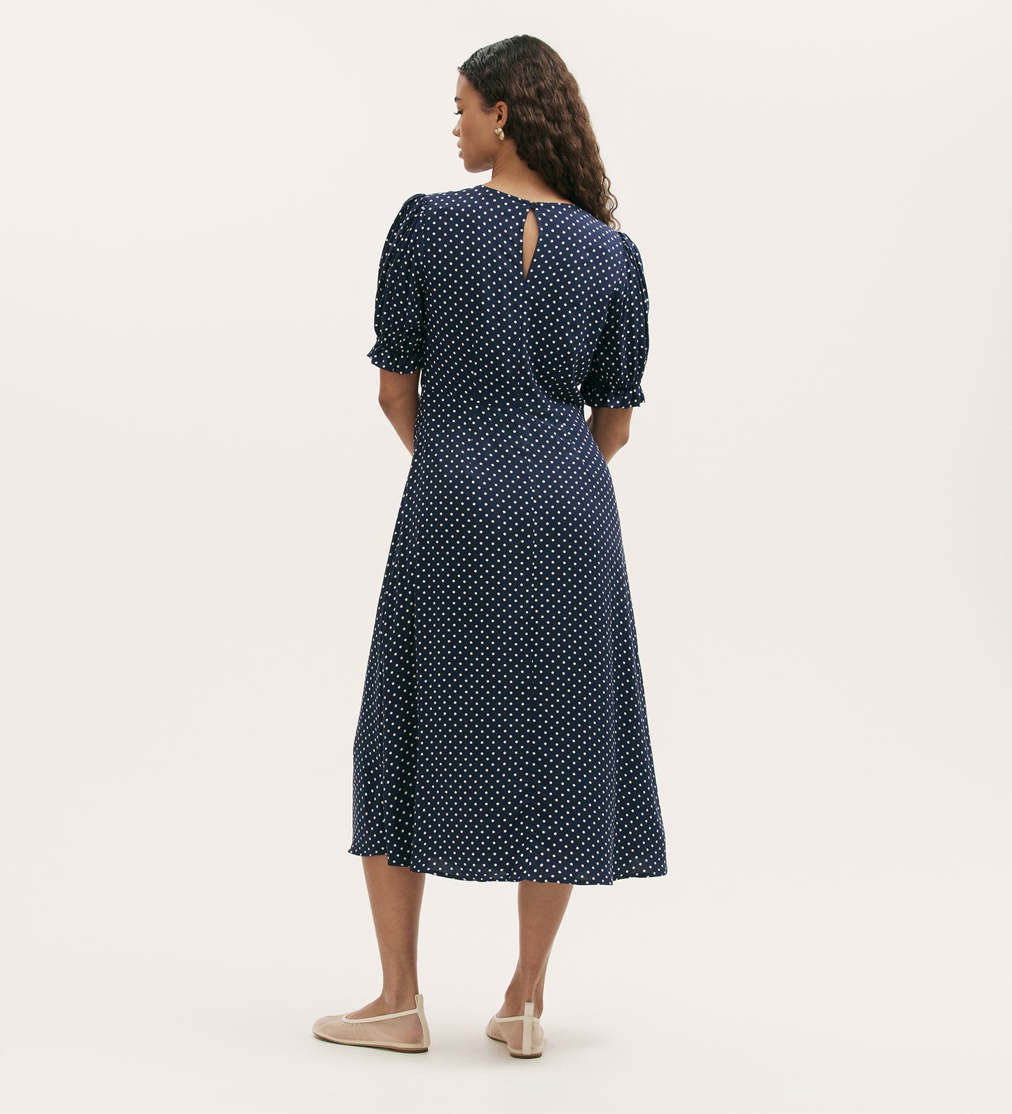 A woman with curly hair stands facing away, wearing the Mela Navy Polka Dot Midi Tea Dress, a chiffon dress with an empire waist, round neckline, and beige flat shoes, set against a plain light background.