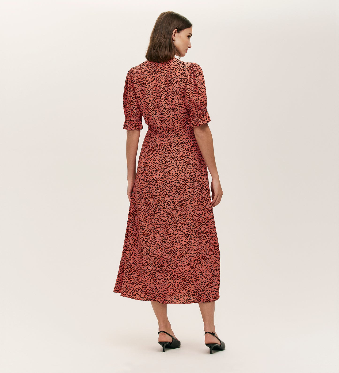 A woman faces away, wearing the Mela Orange Leopard Crepe Midi Tea Dress with short sleeves and a black leopard print. She pairs it with black low-heeled shoes against a plain, light background.