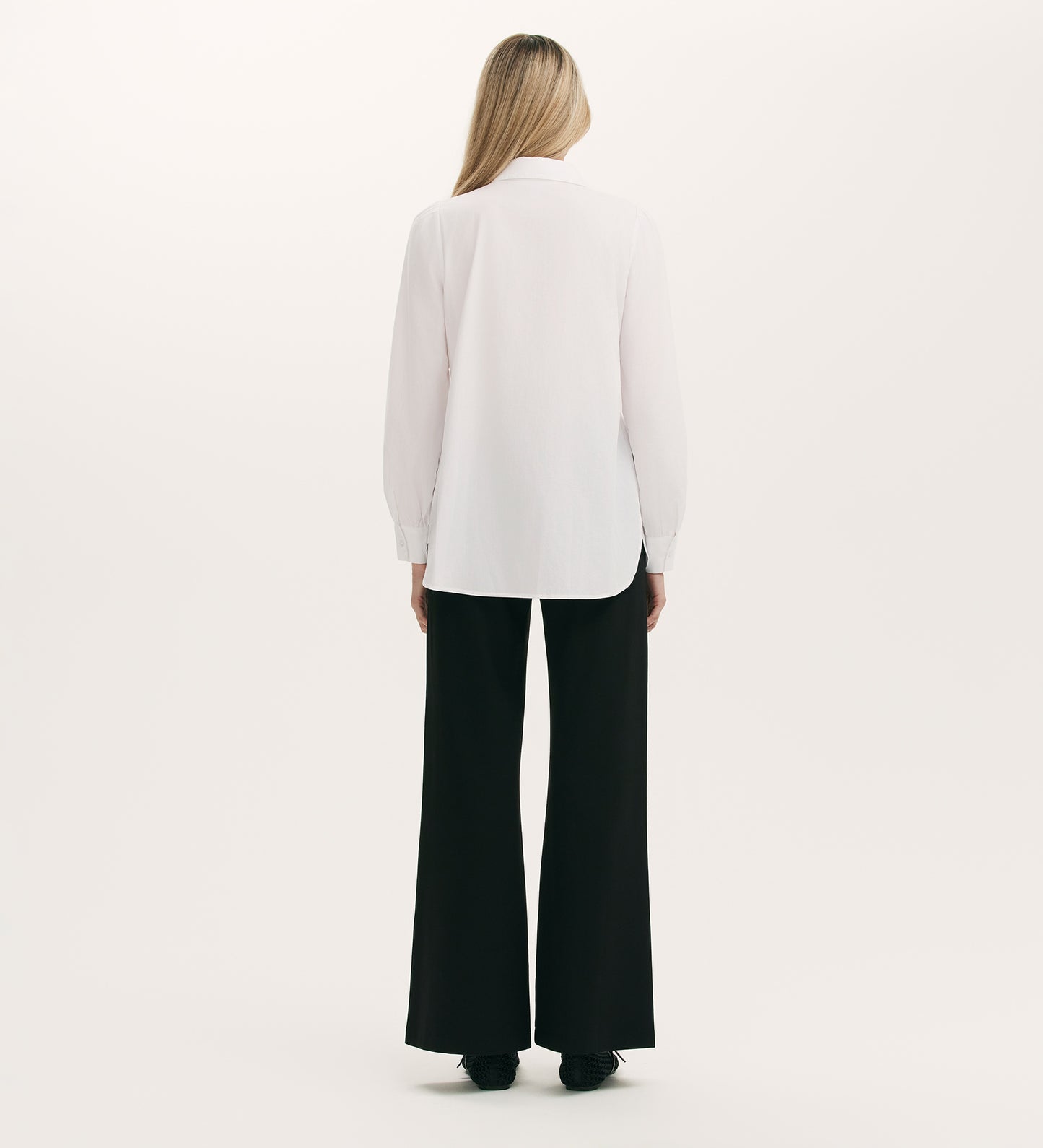 A woman with straight blonde hair stands against a light background, facing away while wearing the Melissa White Cotton Shirt with tailored cuffs and loose black pants paired with black shoes.