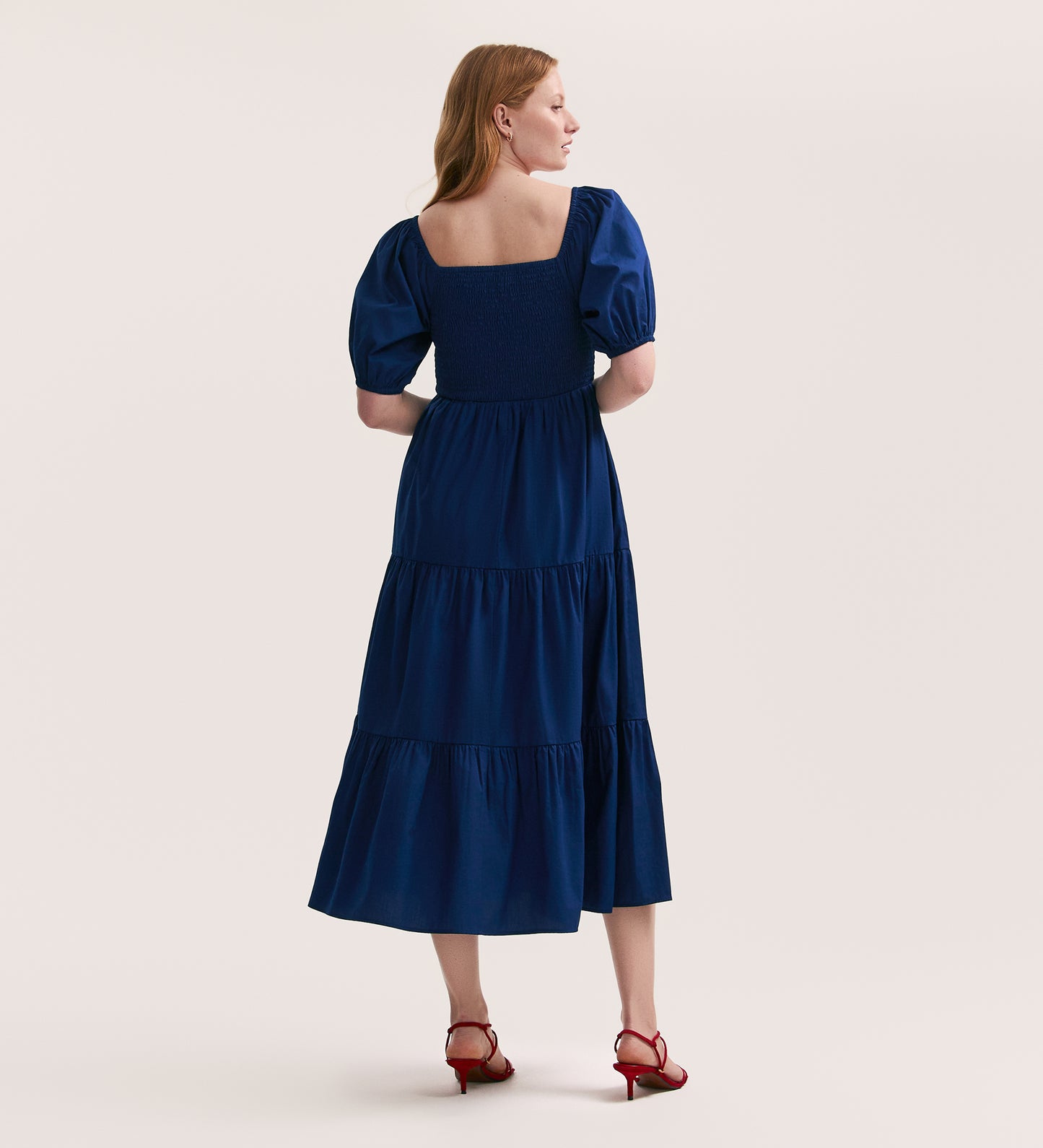 A woman with light skin and long red hair faces away, wearing the Melody Indigo Cotton Midi Tiered Dress with puff sleeves and red heeled sandals, set against a plain light background.