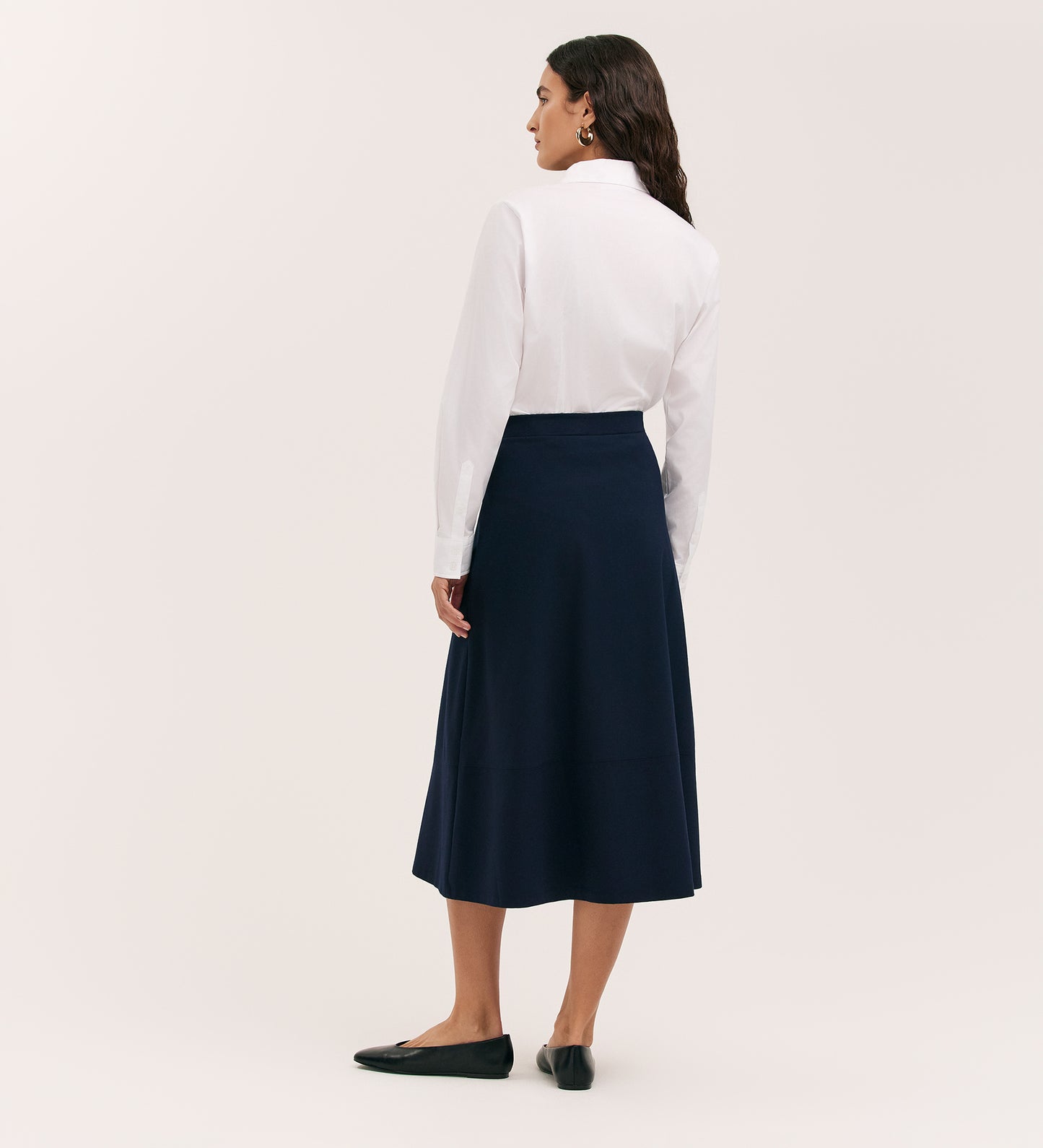 A woman with dark hair stands facing away, wearing a white button-up shirt tucked into the Mikia Navy Ponte Jersey Midi Skirt with an elasticated waistband. She wears black flats and stands against a plain light background.