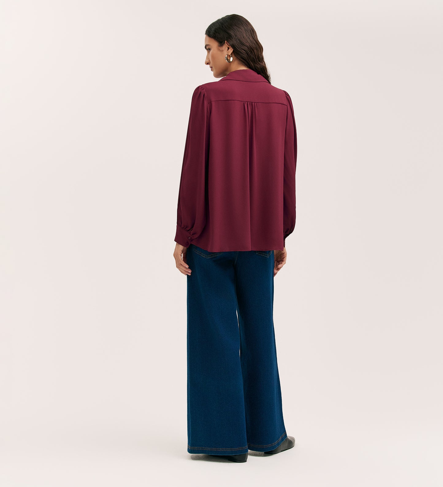 A woman stands facing away in the Melina Burgundy Crepe Blouse, a relaxed fit long-sleeved shirt paired with wide-leg blue jeans against a plain light background.