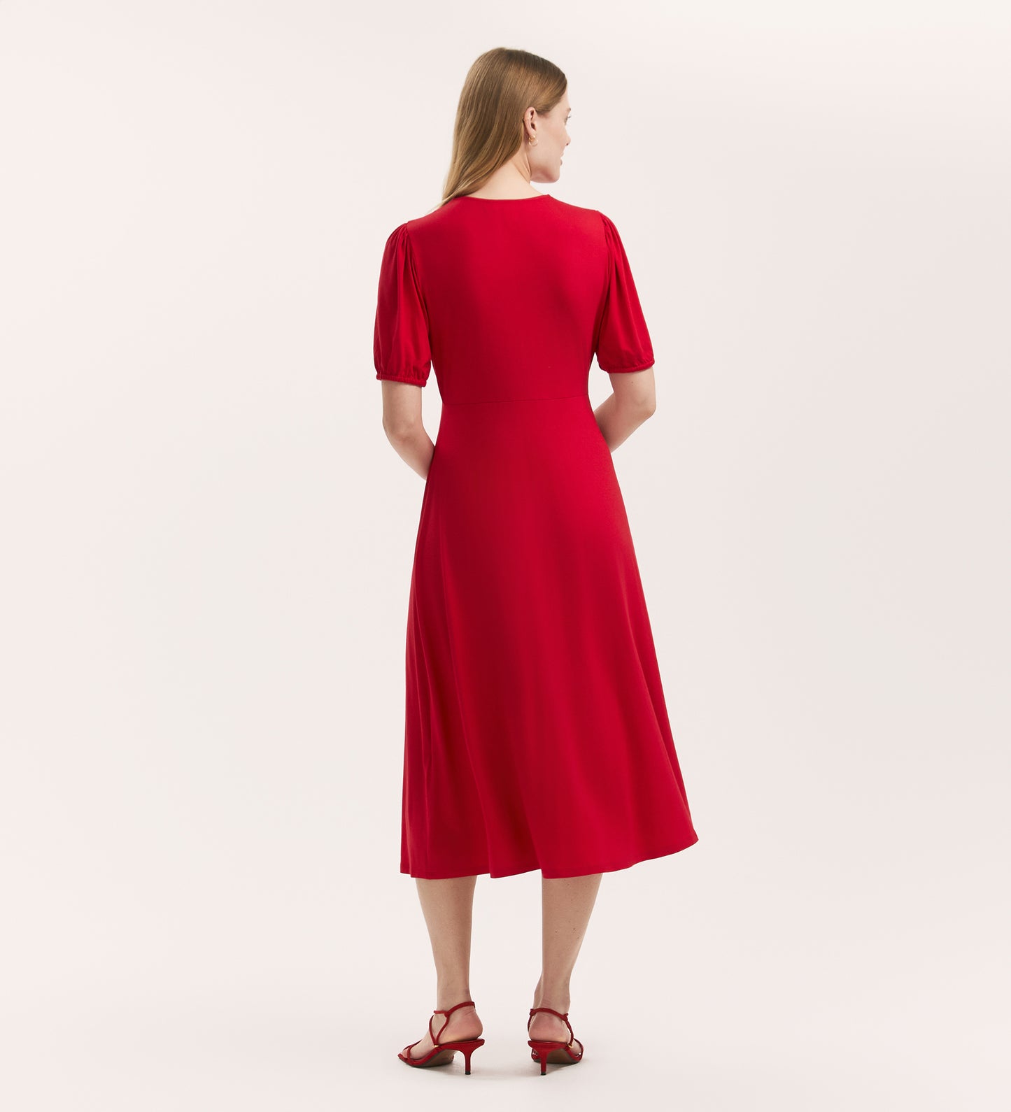 A woman with long blonde hair wears the Milena Red Jersey Button Through Midi Dress and matching red heeled sandals, standing facing away against a plain, light background.