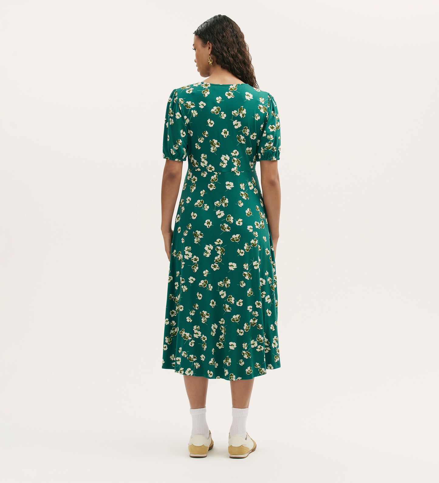 A woman with curly hair stands facing away in the Milena Green Poppies Jersey Midi Dress, featuring a green V-neck, short sleeves, and yellow-and-white floral print. She wears white socks and yellow sneakers against a plain white background.
