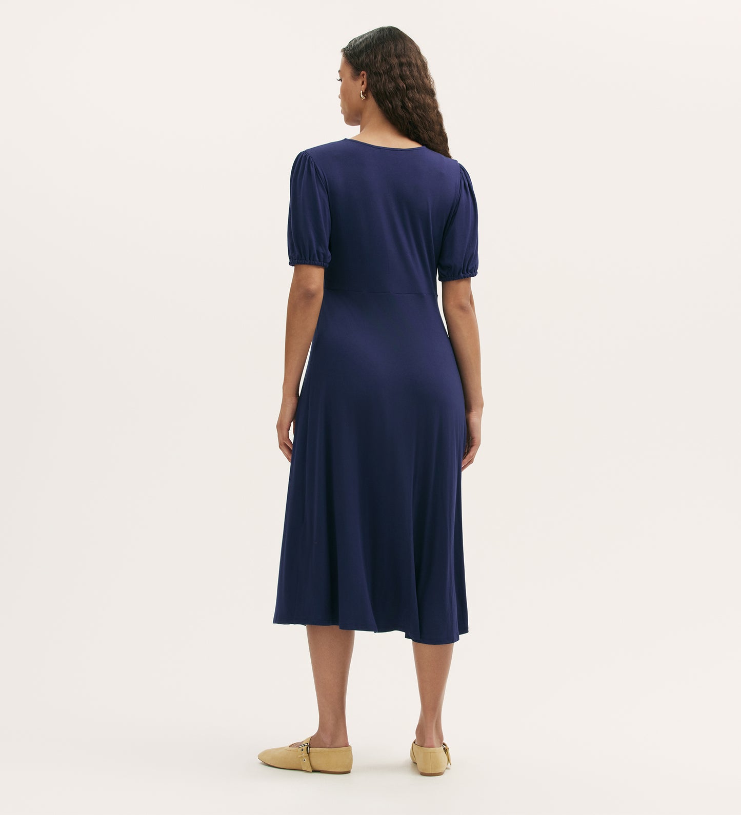 A woman with long wavy hair stands facing away, wearing the Milena Navy Jersey Midi Dress with short puff sleeves and tan flats against a plain light background.