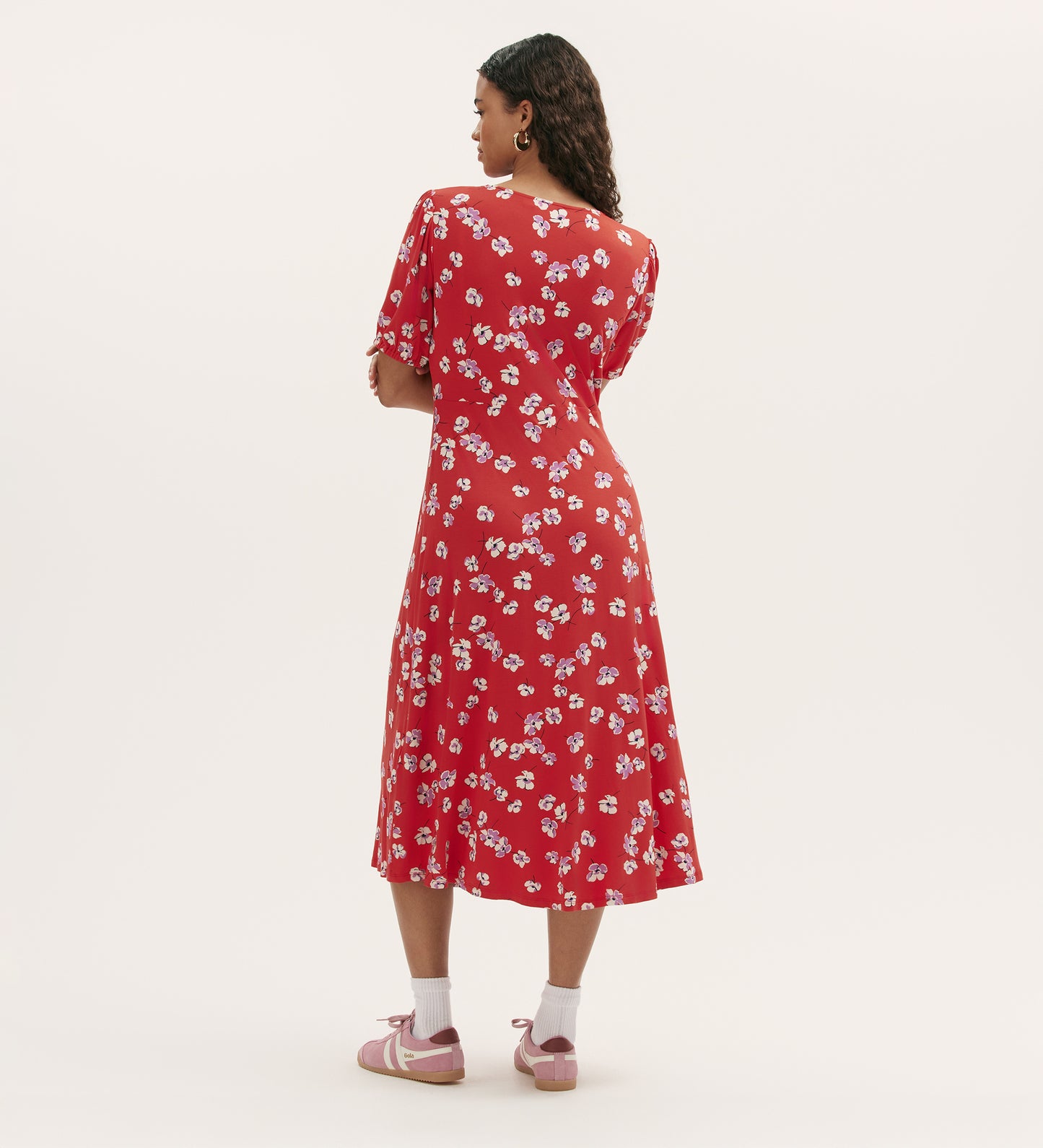 A woman with curly hair stands facing away, wearing the Milena Red Poppies Jersey Midi Dress with a V-neck and short sleeves. She pairs it with white socks and pink sandals against a plain light background.