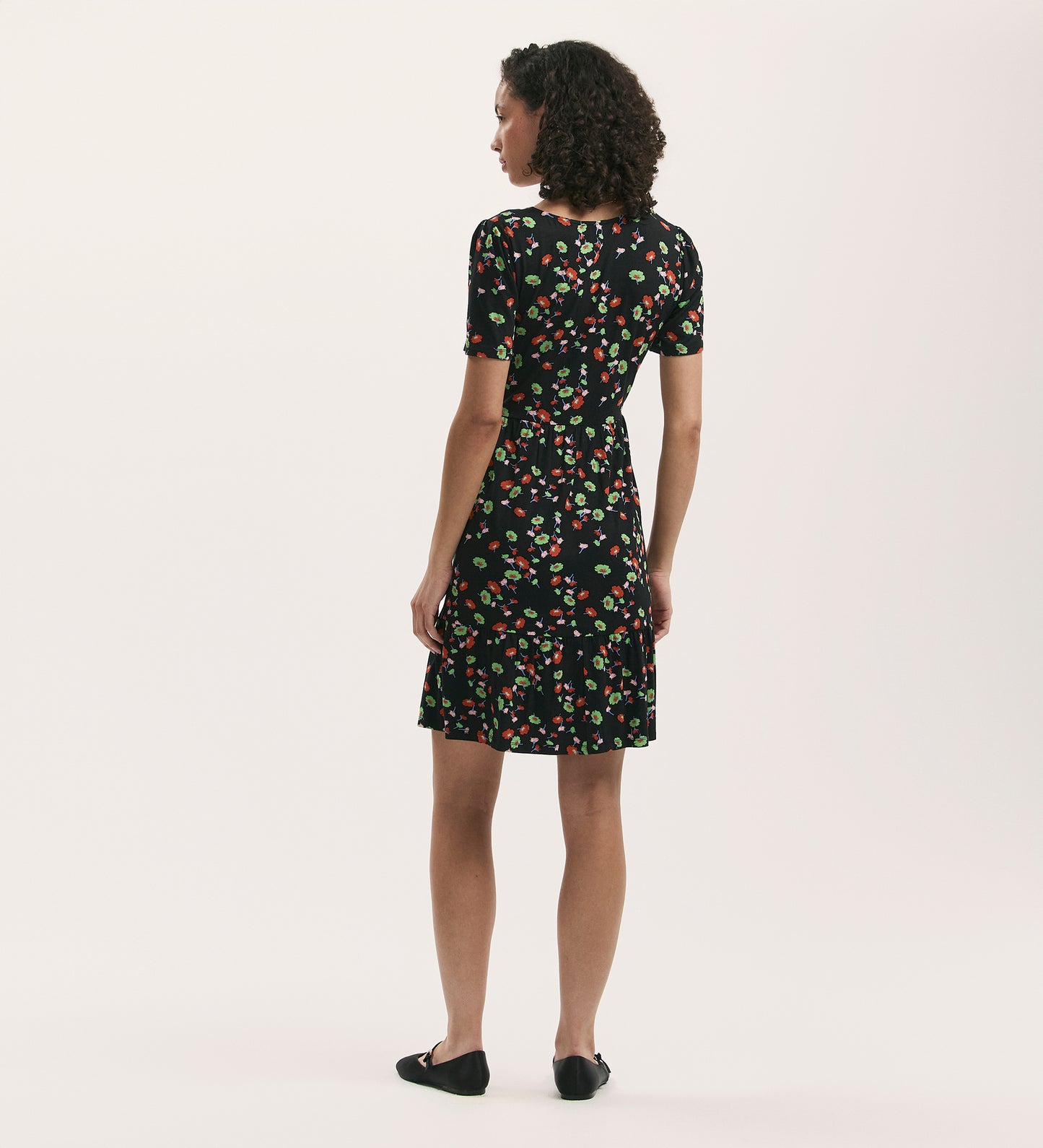 A woman with curly hair faces away, wearing the Misha Black Flower Jersey Short Tiered Dress and black flats, set against a plain light background.