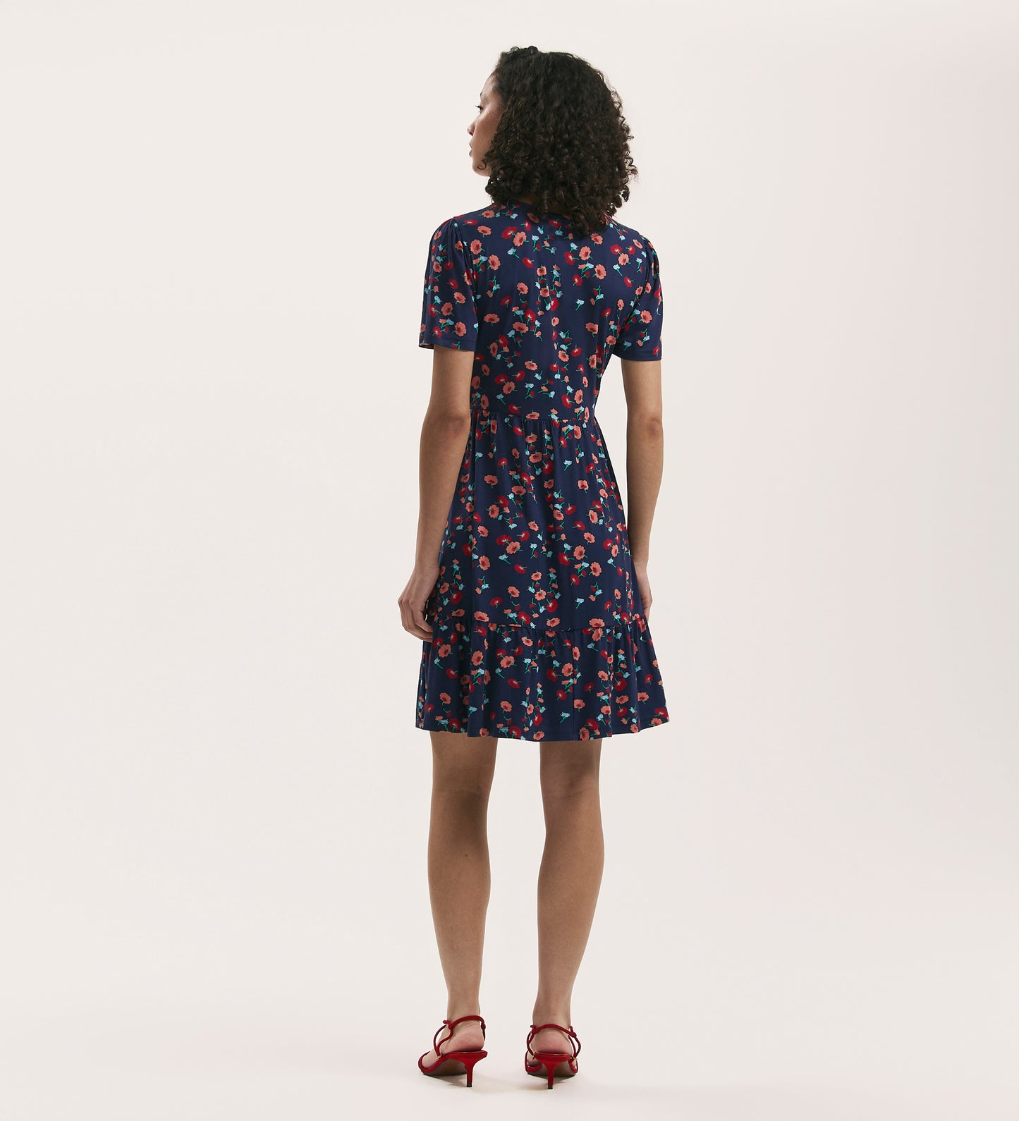 A woman with curly hair faces away, wearing the Misha Navy Flower Jersey Short Tiered Dress with a v-neck and tiered skirt, paired with red heeled sandals, set against a plain light background.