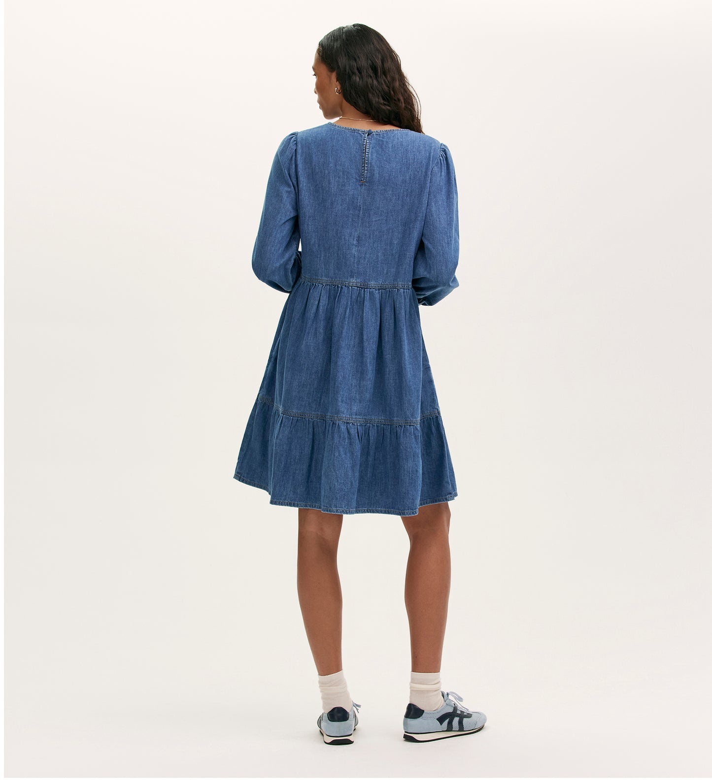 A woman faces away wearing the Nancy Light Blue Denim Dress, a relaxed fit with long sleeves and a tiered skirt, styled with white socks and blue sneakers against a neutral background.