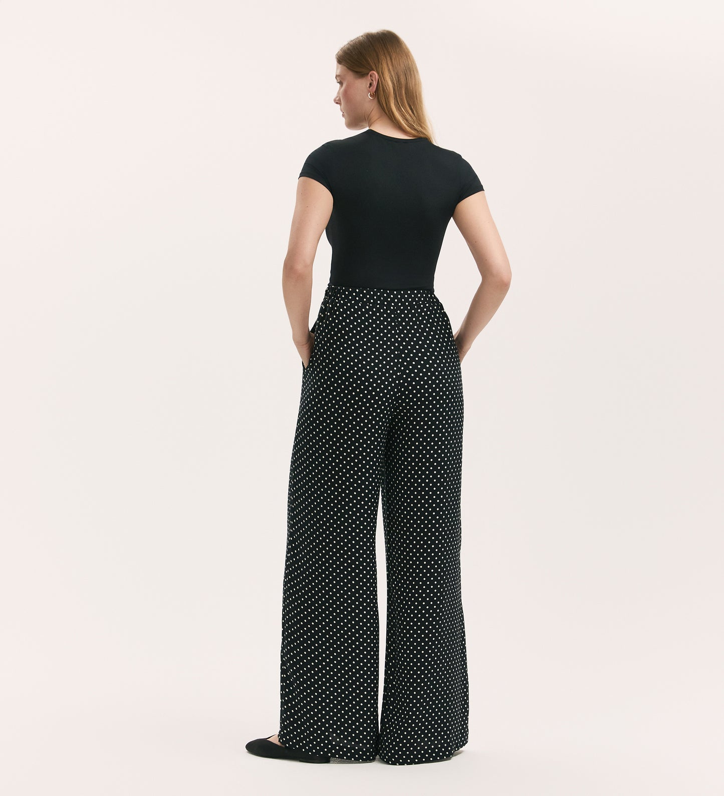 A woman stands with her back to the camera, wearing a fitted black short-sleeve top and Nat Black Polka Dot Trousers—high-waisted, wide-leg palazzo pants with a white polka dot pattern—her hands resting on her hips against a plain background.
