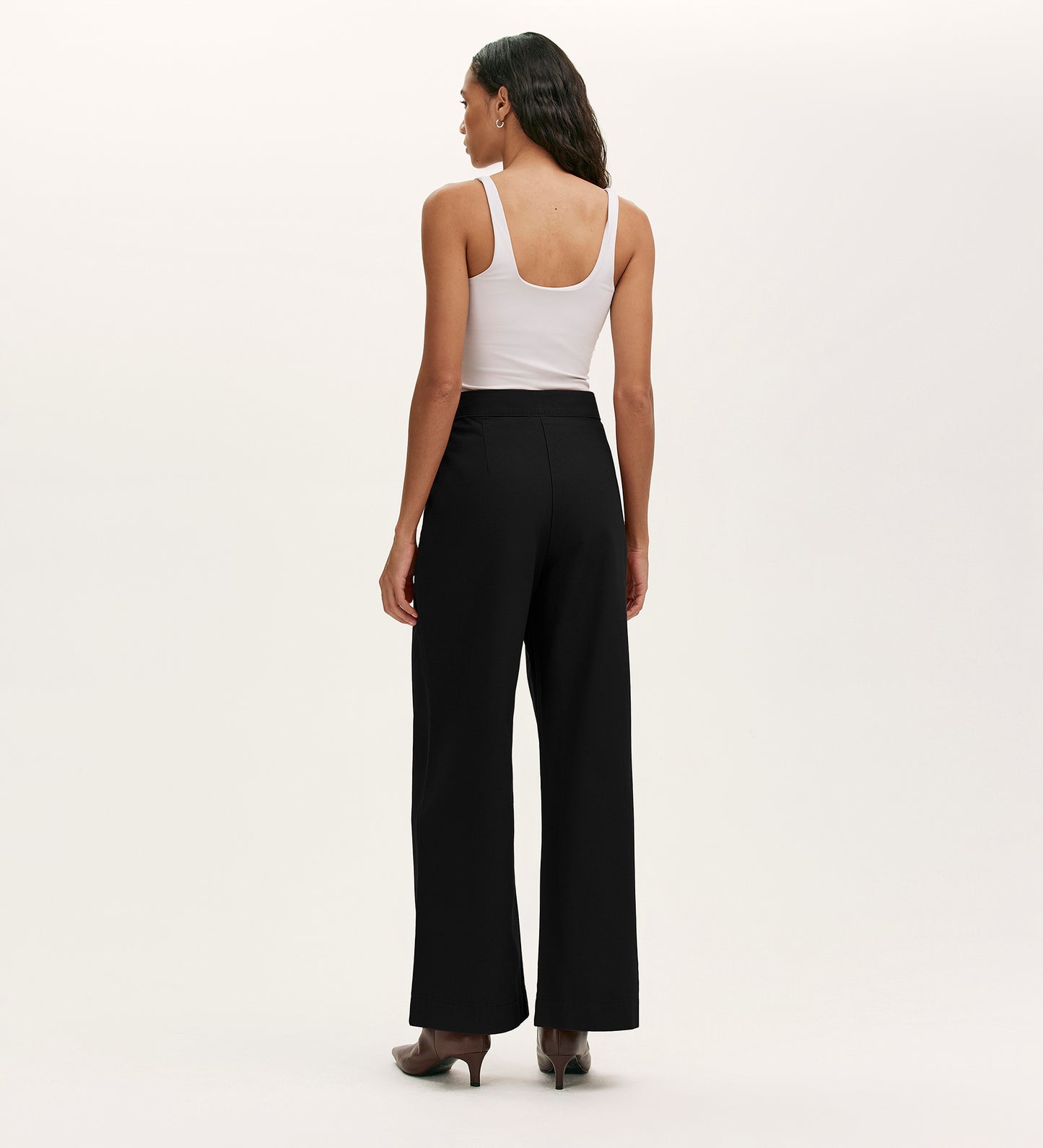 A woman faces away, wearing a white sleeveless top, heeled shoes, and the Nellie Black Canvas Wide Leg Trousers—a versatile wardrobe staple—against a plain, light-colored background.