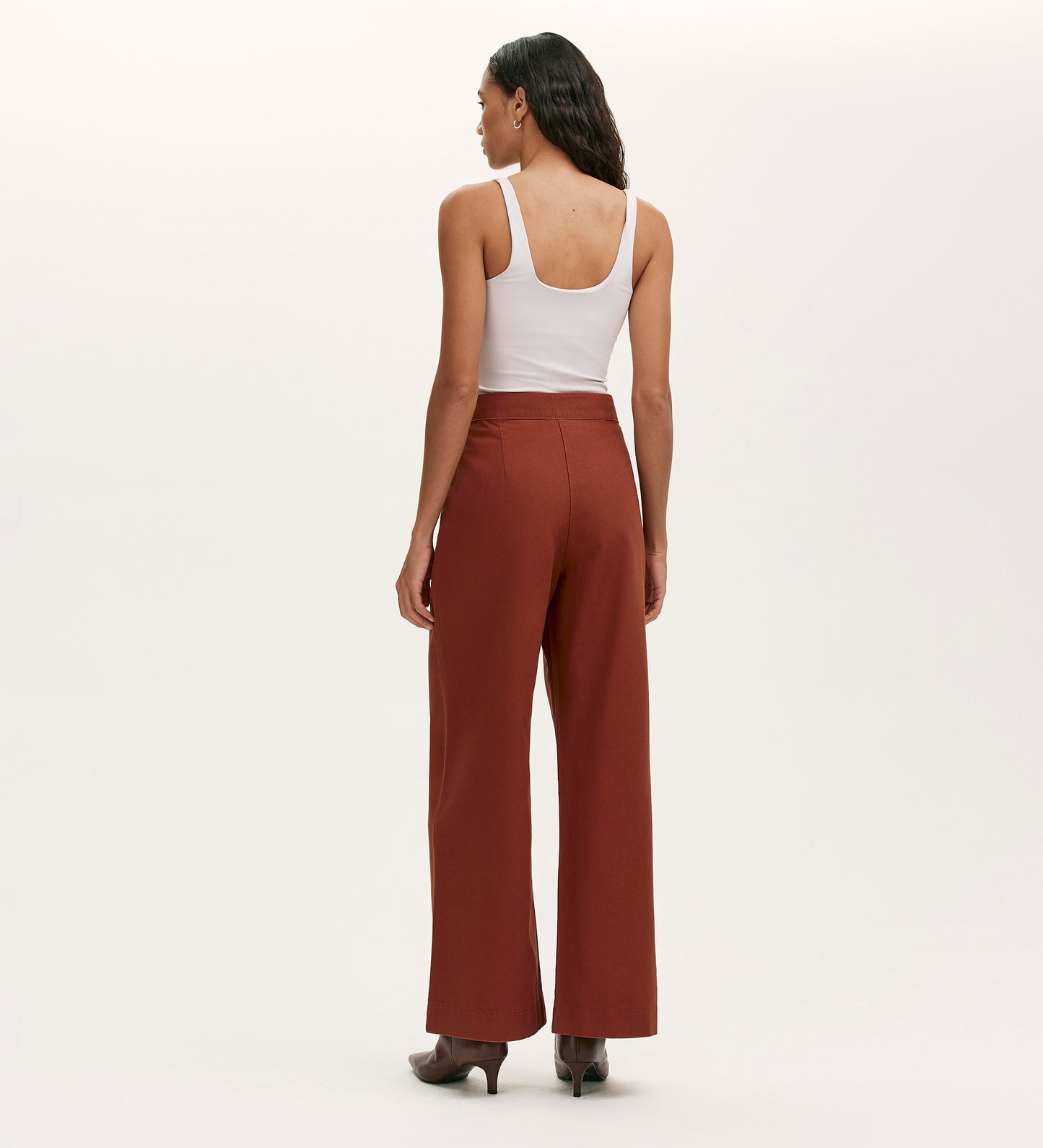 A woman stands facing away, wearing a white sleeveless top, Nellie Terracotta Canvas Wide Leg Trousers, and heeled boots against a plain light background.