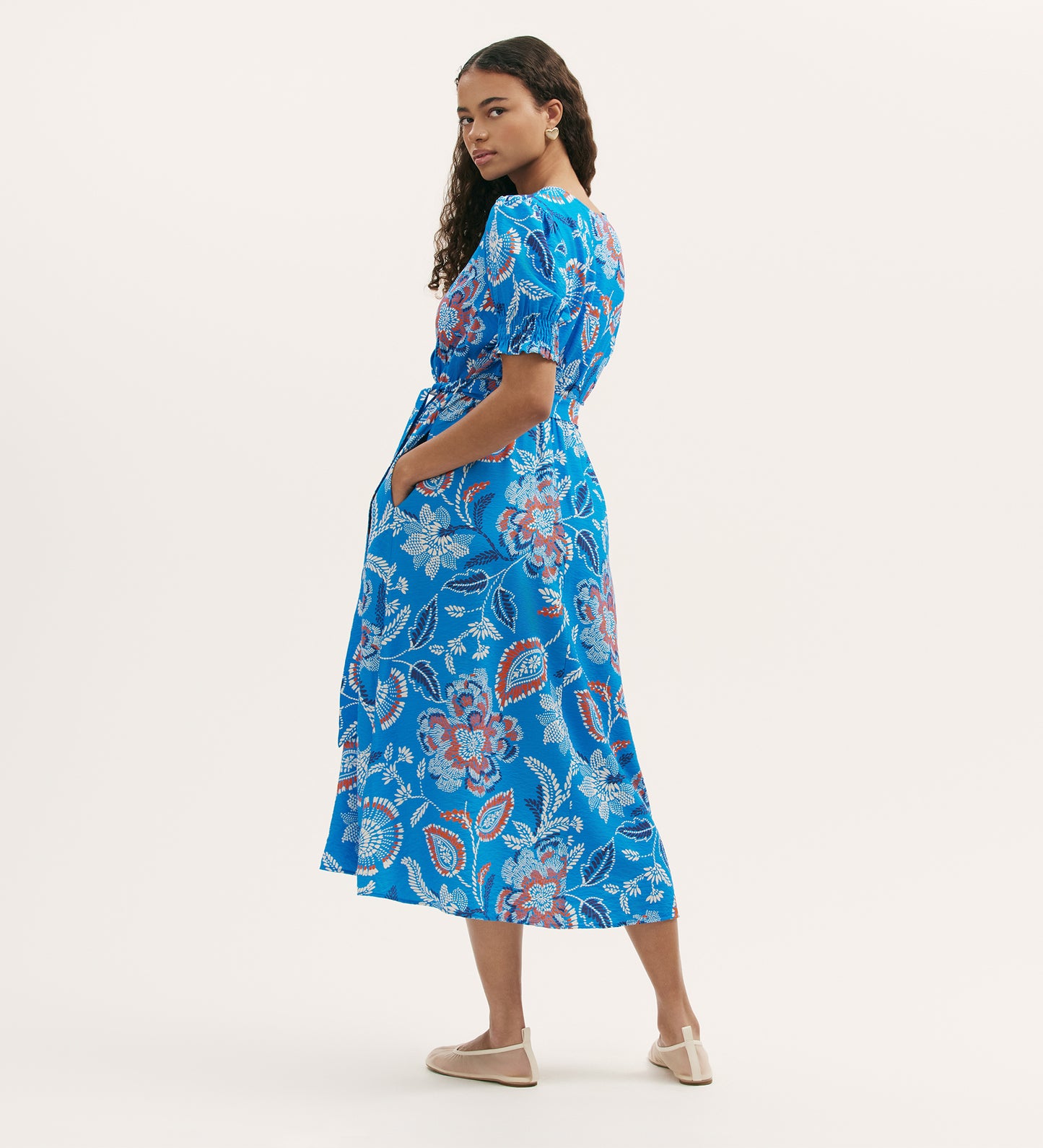 A woman with long curly hair stands sideways against a plain light background, hands in her pockets, wearing the Nicoleta Blue Floral Midi Dress with a V-neck, short sleeves, shirred waist, and beige flats.