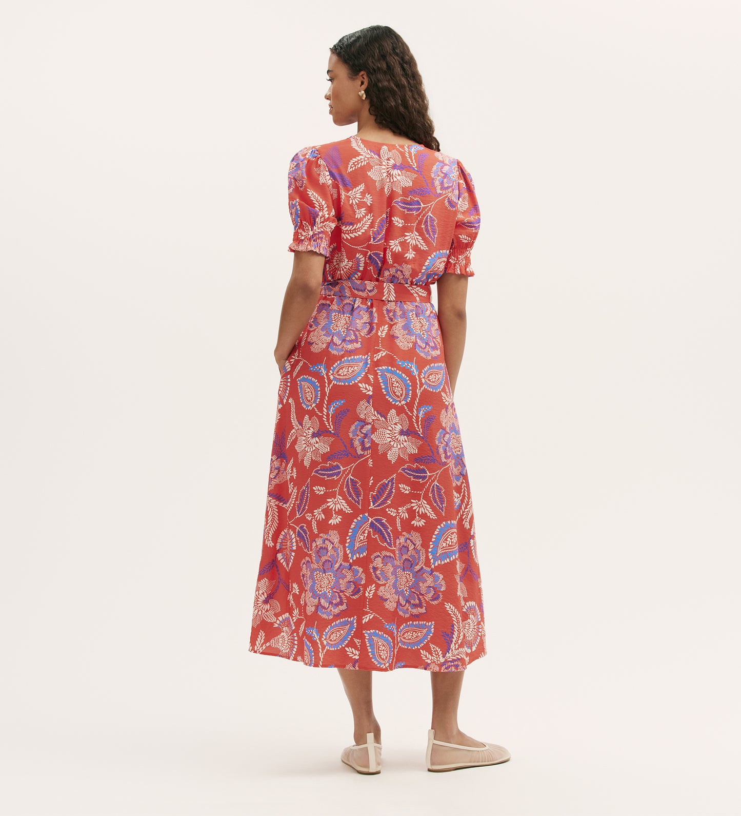 A woman stands with her back to the camera, wearing the Nicoleta Coral Floral Midi Dress—featuring a red floral print, V-neck, puff sleeves, and shirred waist—paired with beige flats against a plain light background.