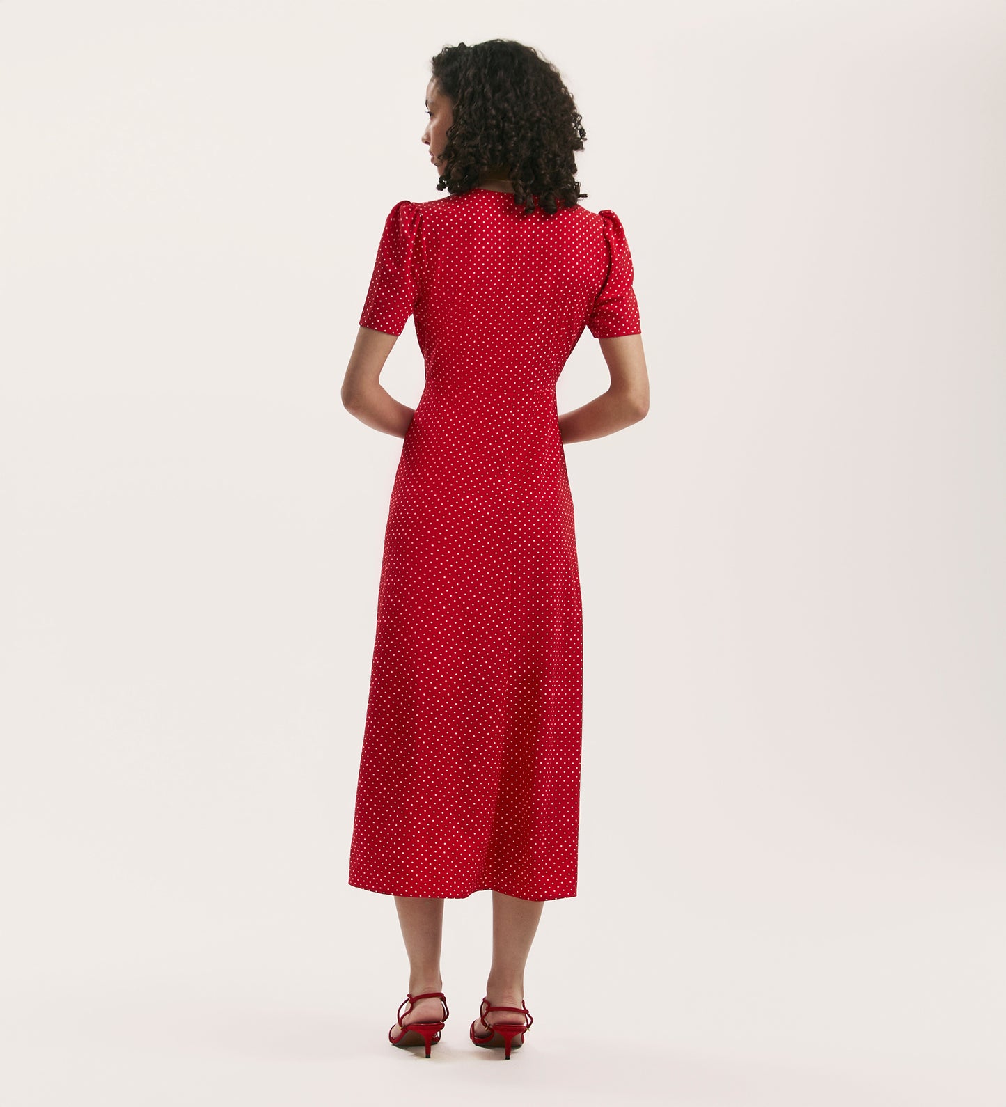 A woman, seen from behind against a plain light background, wears the Nieve Red Polka Dot Crepe Midi Tea Dress paired with red heeled sandals.