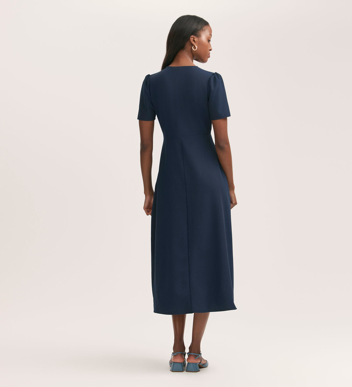 A woman faces away, wearing the Nieve Navy Crepe Midi Tea Dress with button details and blue heeled sandals, set against a plain light background.