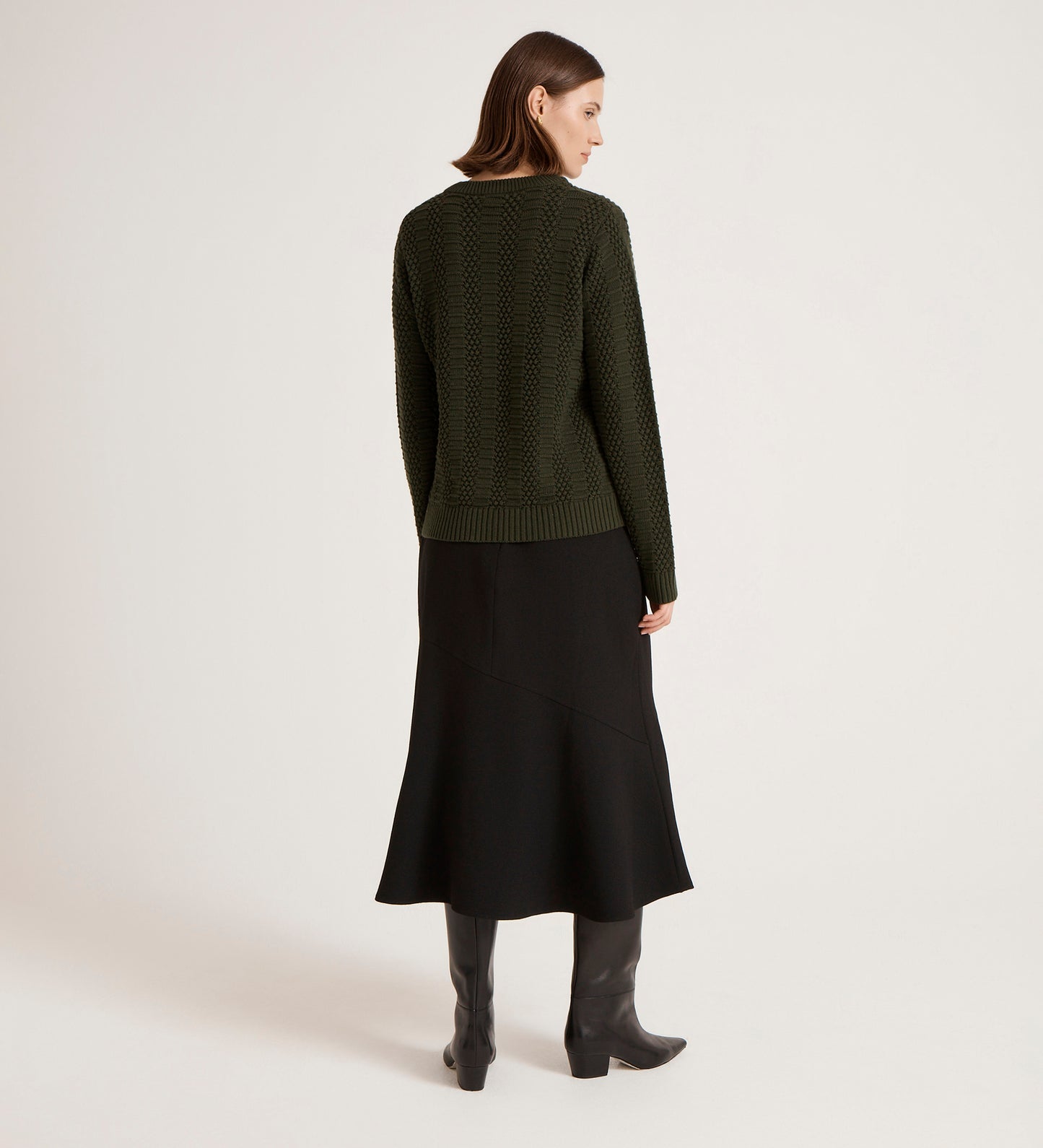 A woman stands facing away, wearing the Nova Green Knitted Jumper with a black midi skirt and black knee-high boots, against a plain light background.