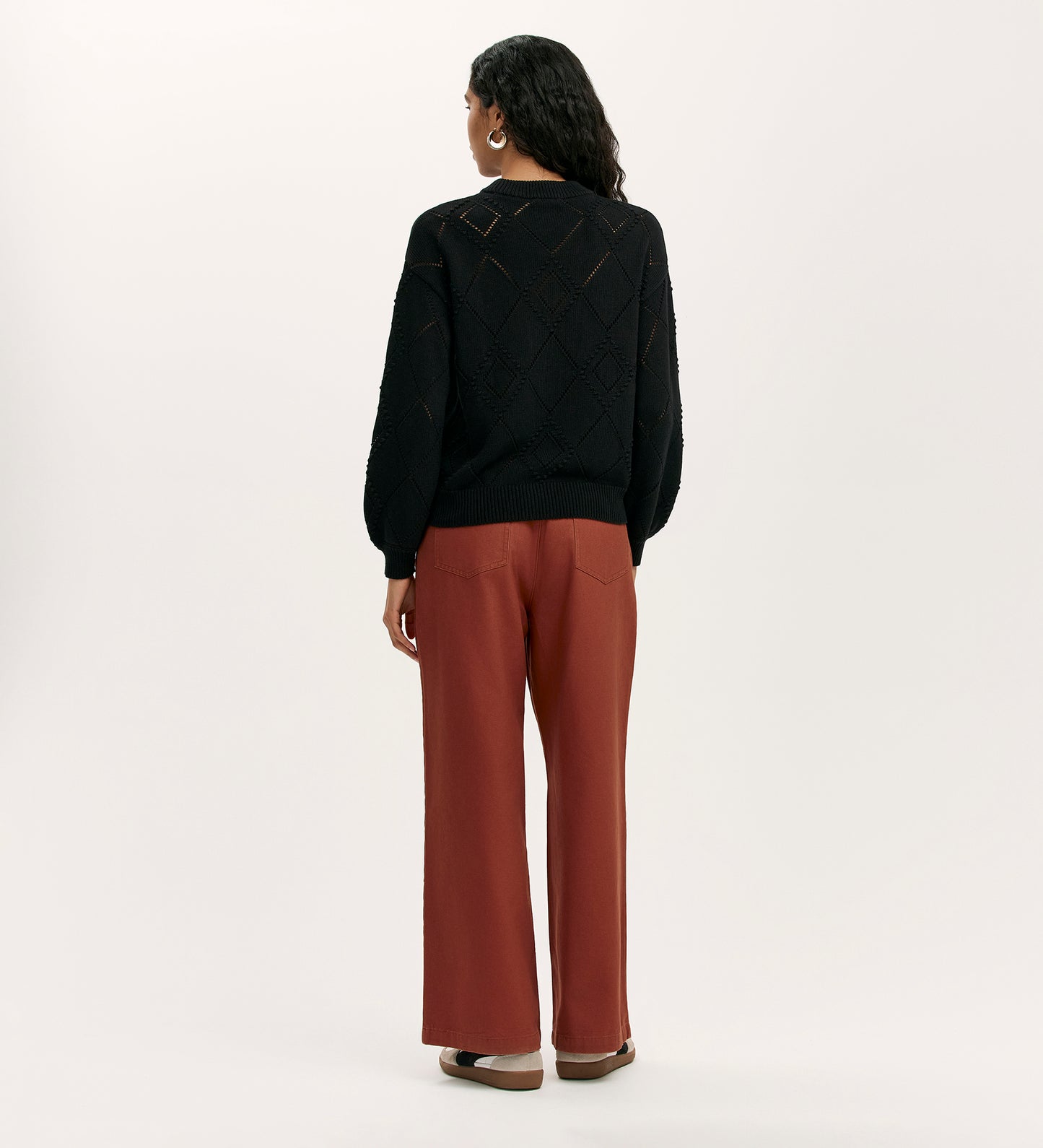 A person with long dark hair, seen from behind, wears the Odette Black Knitted Jumper with ribbed detailing, rust wide-leg pants, and brown shoes against a plain light background.
