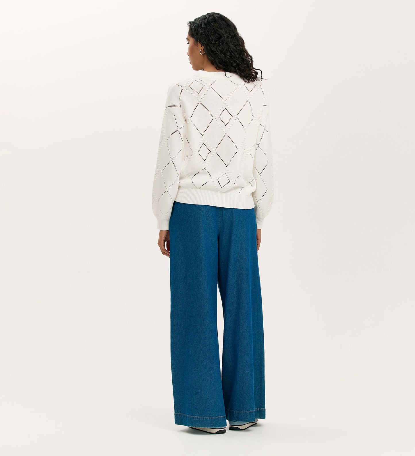 A person with long, curly hair stands facing away against a plain light background, wearing the Odette Ivory Knitted Jumper with a diamond pointelle knit and wide-leg blue jeans.