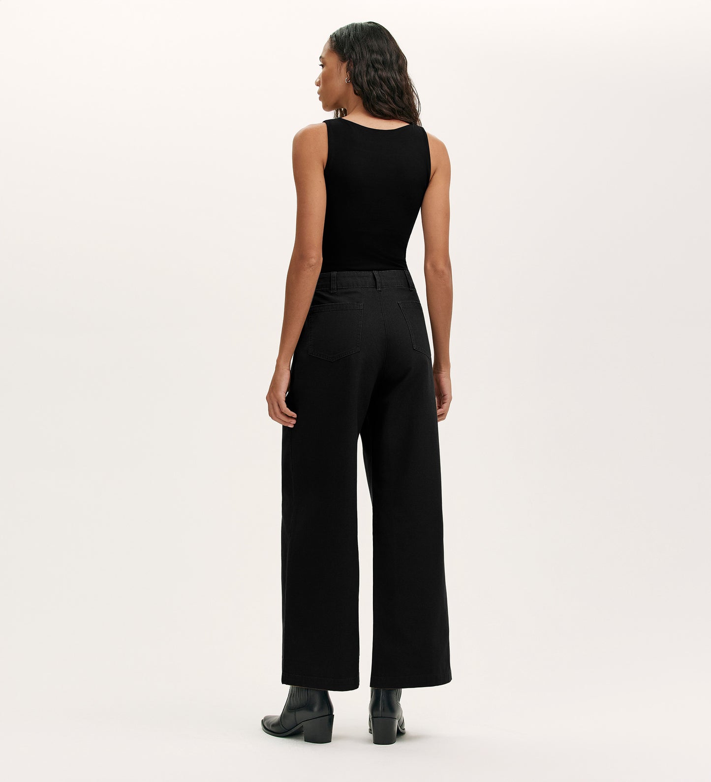 A woman faces away from the camera, wearing Olive Black Canvas Wide Leg Trousers with a sleeveless black top and ankle boots against a plain light background—an effortless elevated casual look.