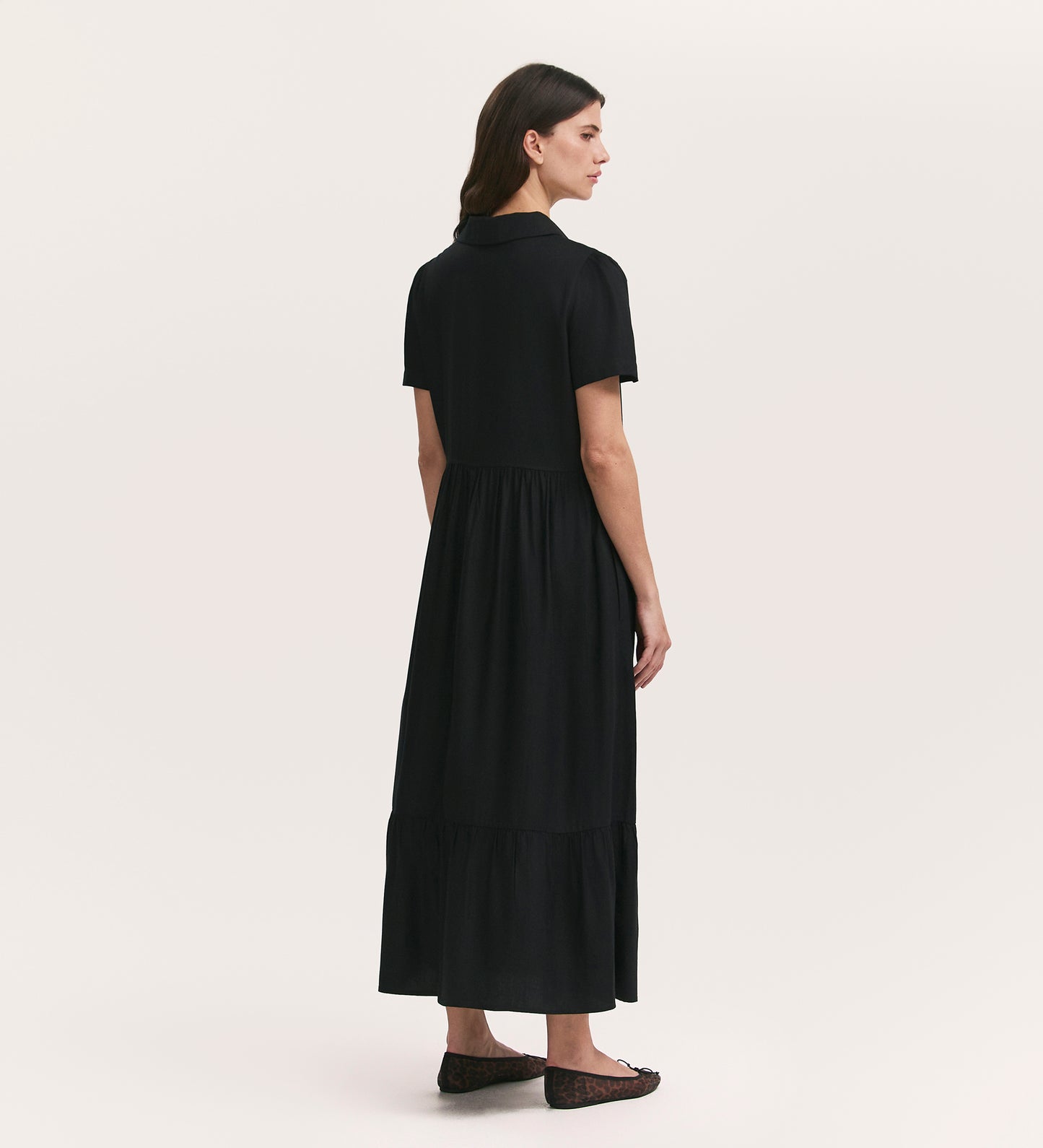 A woman faces away, wearing the Orion Black Zip Front Midi Shirt Dress with a relaxed fit, short sleeves, gathered waist, and flat shoes. The background is plain and light-colored.