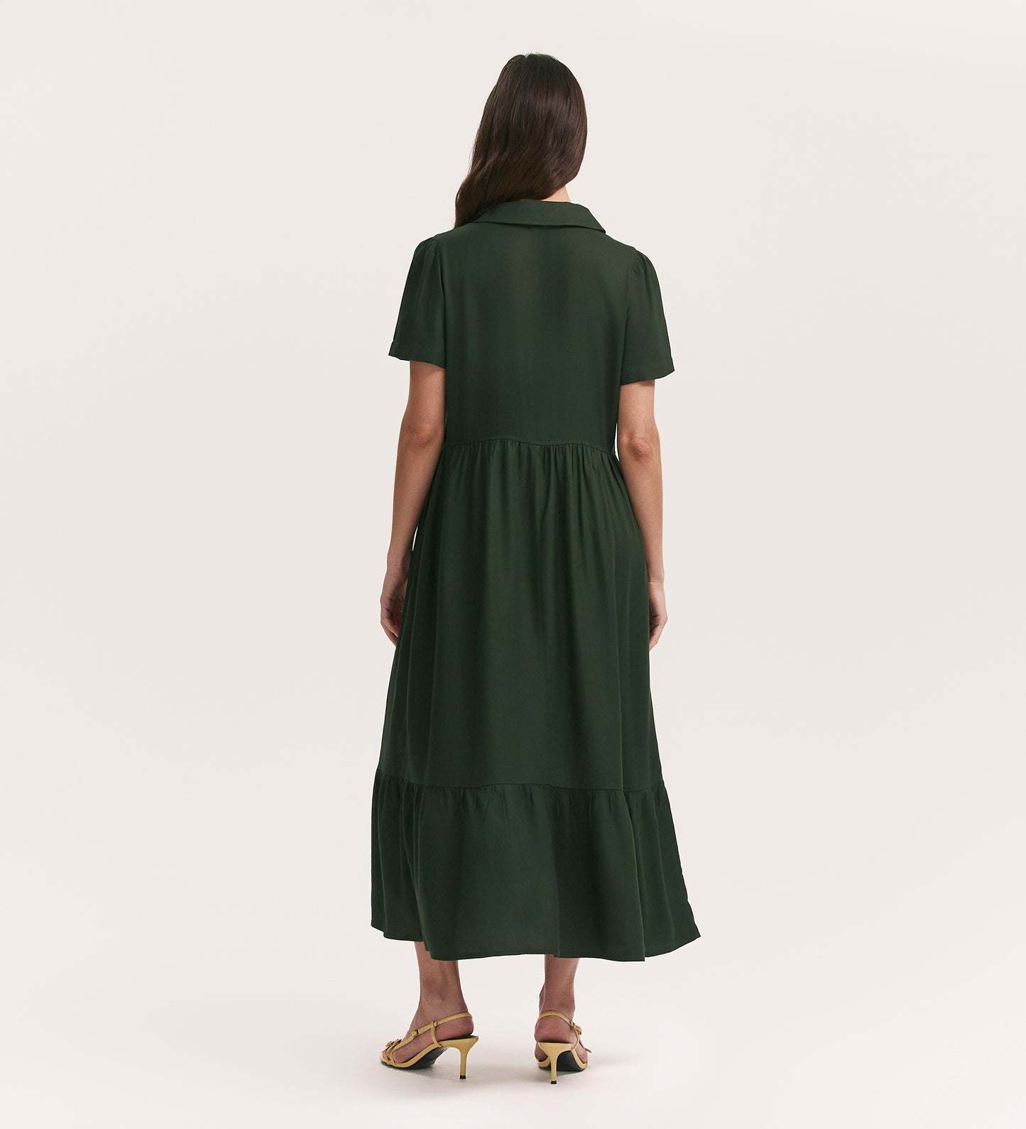 A woman with long brown hair faces away, wearing the Orion Dark Khaki Zip Front Midi Shirt Dress featuring a tiered hemline, short sleeves, functional pockets, and yellow heeled sandals against a plain light backdrop.
