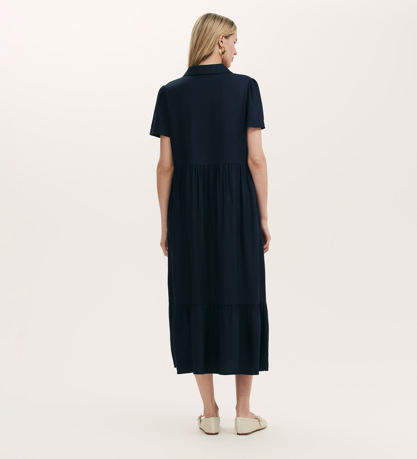 A woman with blonde hair faces away, wearing the Orion Navy Zip Front Midi Shirt Dress featuring short sleeves, a collared neckline, a gathered skirt, and pockets. She pairs it with light flats against a plain light background.