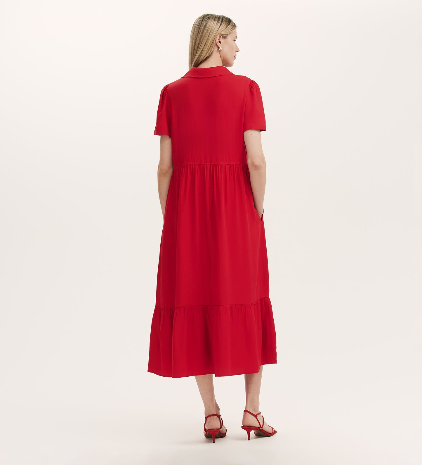 A woman stands facing away, wearing the Orion Red Zip Front Midi Shirt Dress with a relaxed fit, short sleeves, and tiered hemline, paired with red heeled sandals. The background is plain and light-colored.
