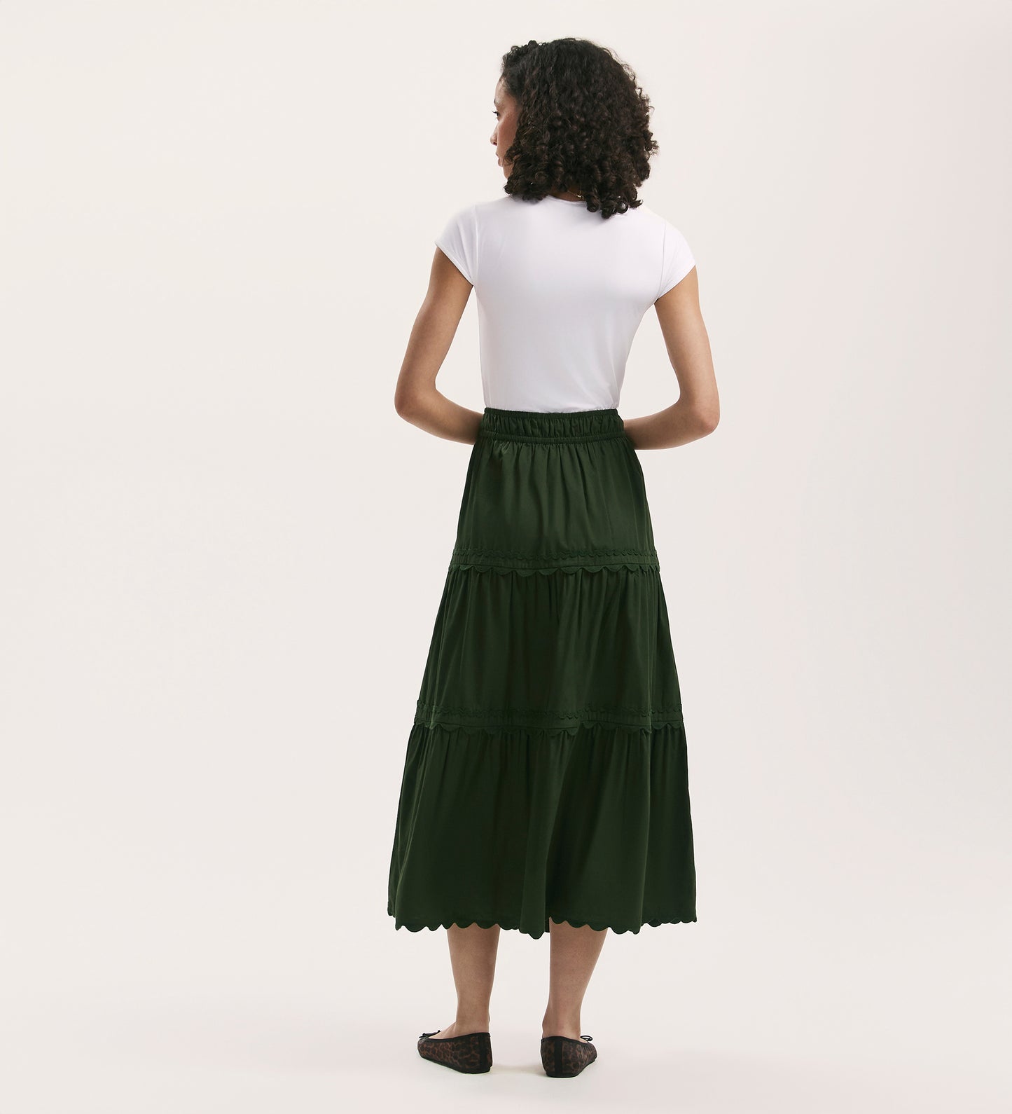 A woman with curly hair, wearing a white short-sleeve top and the Paula Dark Khaki Cotton Ric Rac Trim Midi Skirt, stands facing away from the camera against a plain light background.