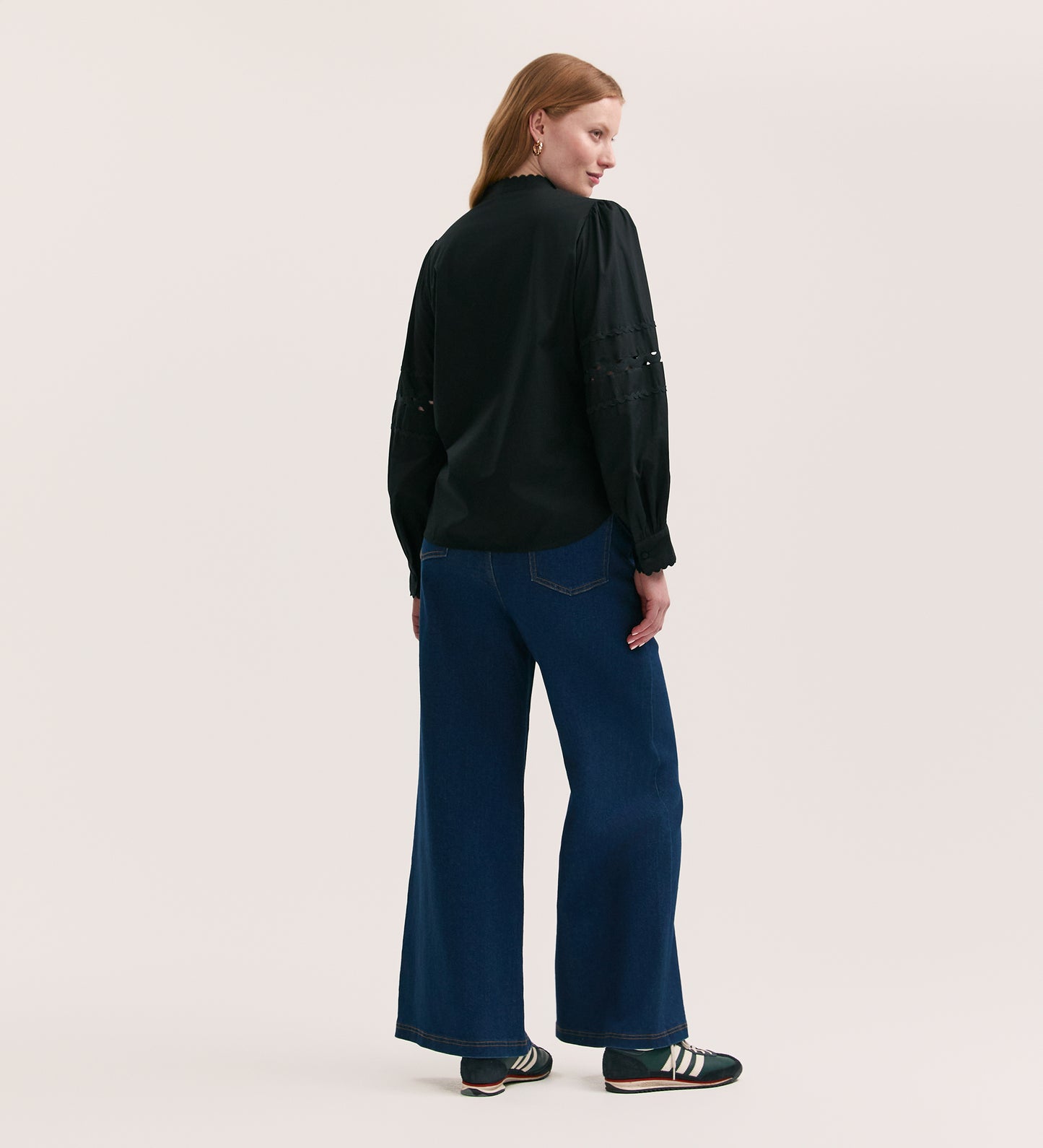 A person with long light brown hair faces away, wearing the Paulie Black Cotton Shirt, wide-leg blue jeans, and black sneakers with white stripes against a plain light background.