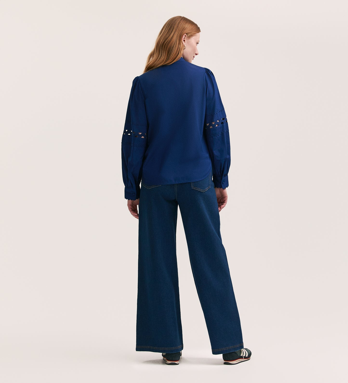 A woman stands with her back to the camera, wearing the Paulie Indigo Cotton Shirt—a relaxed-fit navy blouse with cut-out sleeve details—paired with wide-leg dark blue jeans and black shoes against a plain light background.