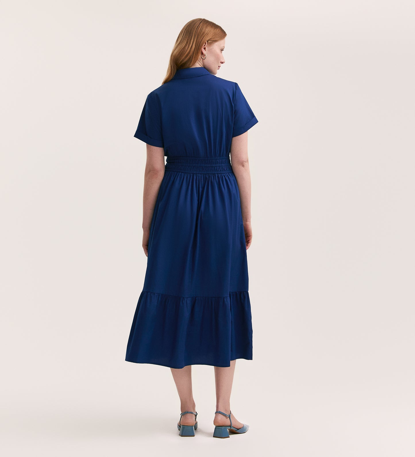 A woman with light brown hair wears the Penny Indigo Cotton Midi Shirt Dress and blue-heeled shoes, standing with her back to a plain light background.