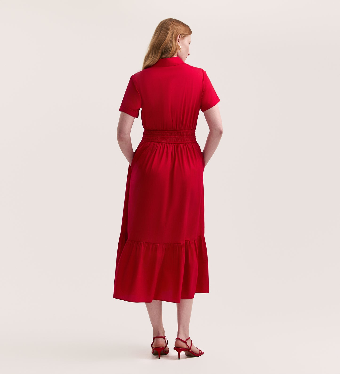 A woman with long red hair stands facing away, wearing the Penny Red Midi Dress featuring an elasticated waist and ruffled hem, paired with matching heeled sandals. The plain light background highlights her chic look.