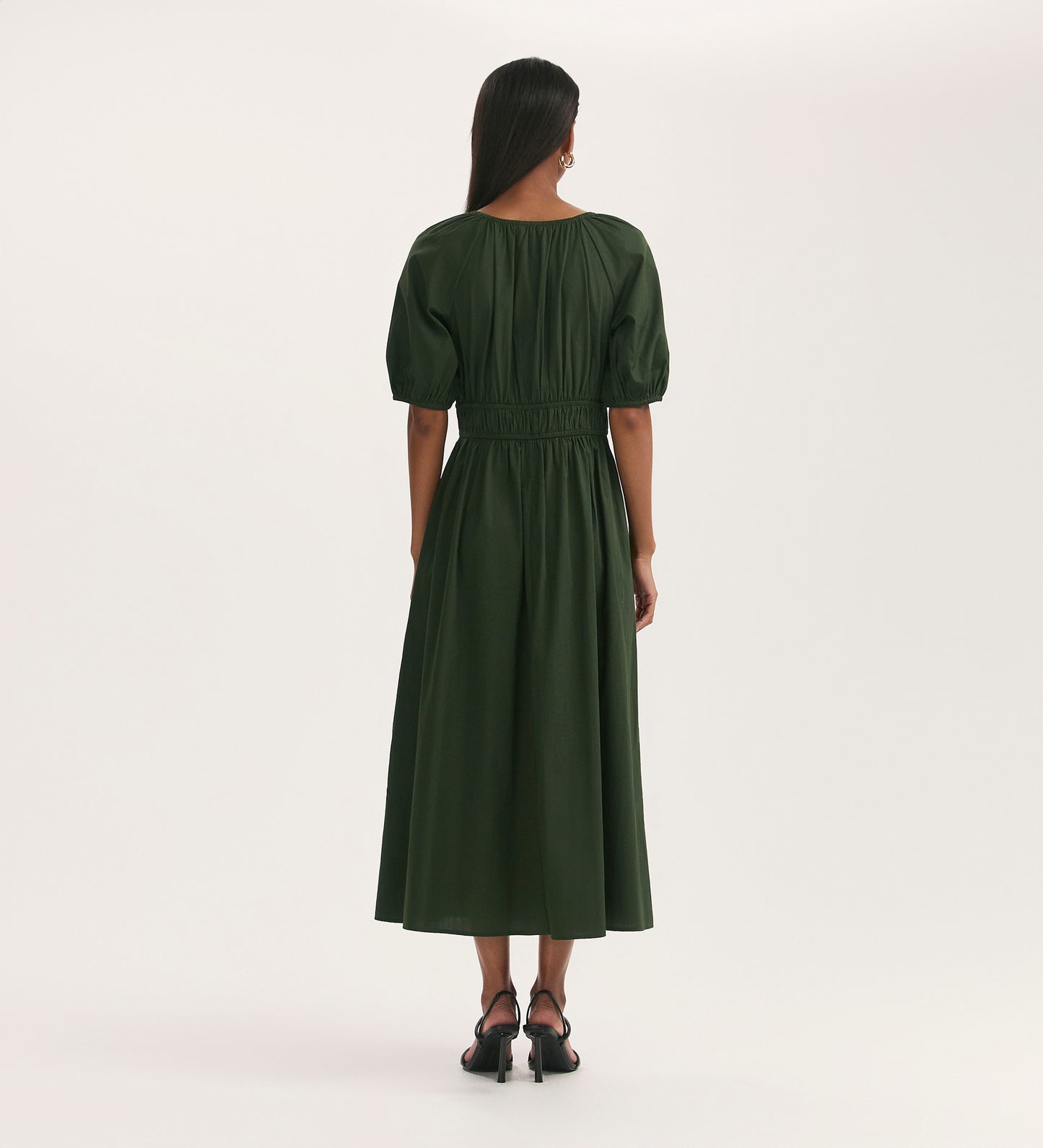 A woman with long dark hair, facing away, wears the Piper Dark Khaki Cotton Midi Smock Dress featuring a shirred waist. She pairs it with black heeled sandals against a plain light background.
