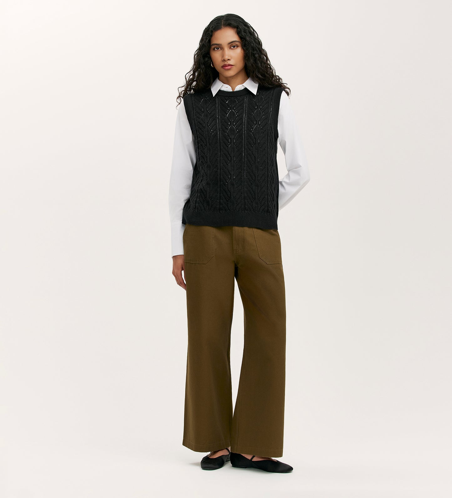 Wearing the Prim Black Knitted Vest over a white collared shirt, paired with brown wide-leg pants and black flats, a person with long curly hair stands against a plain light background.