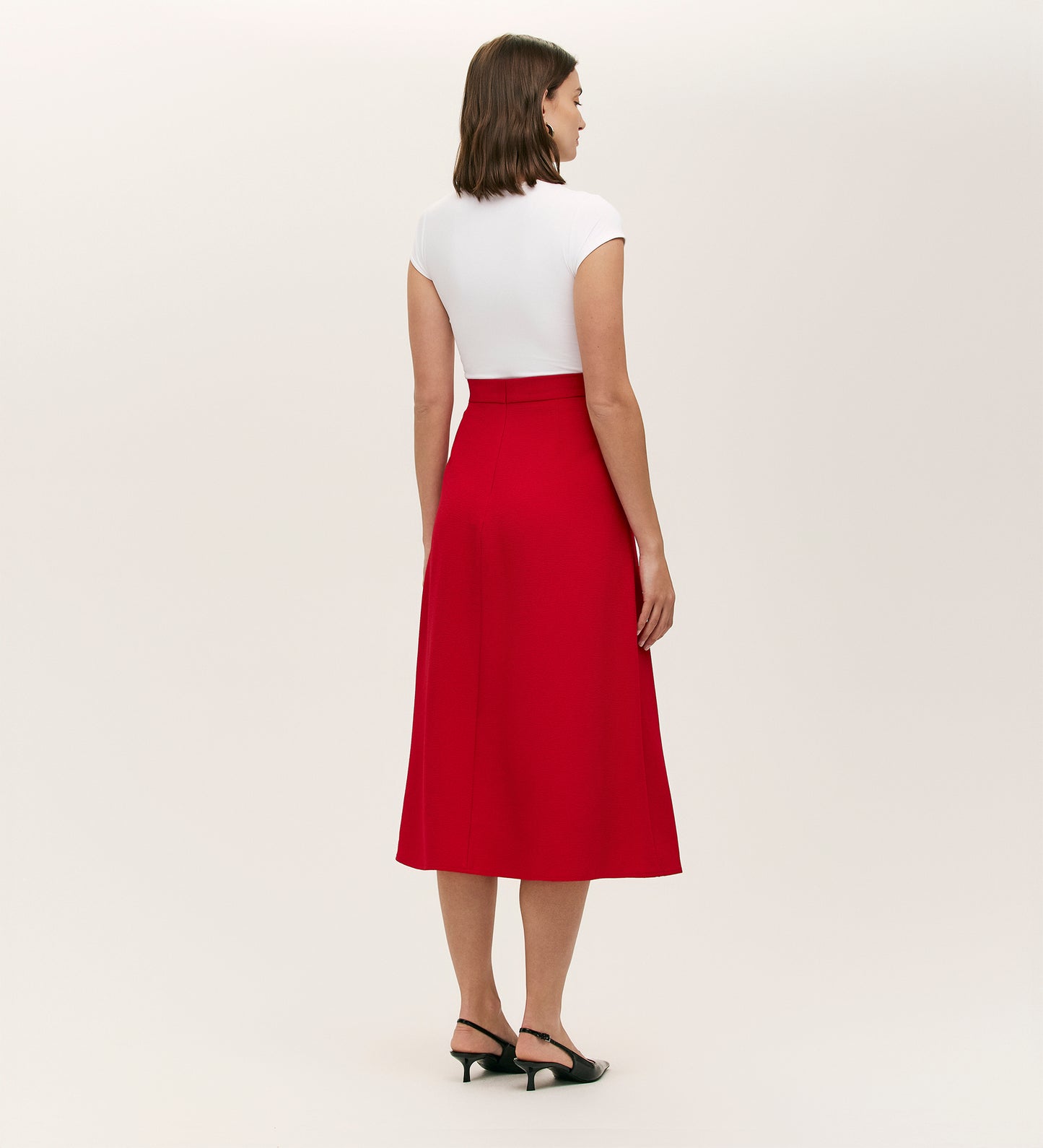 A woman faces away, wearing a white short-sleeve top, black heeled sandals, and the Ramona Red Rich Crepe Midi Skirt against a plain light background.