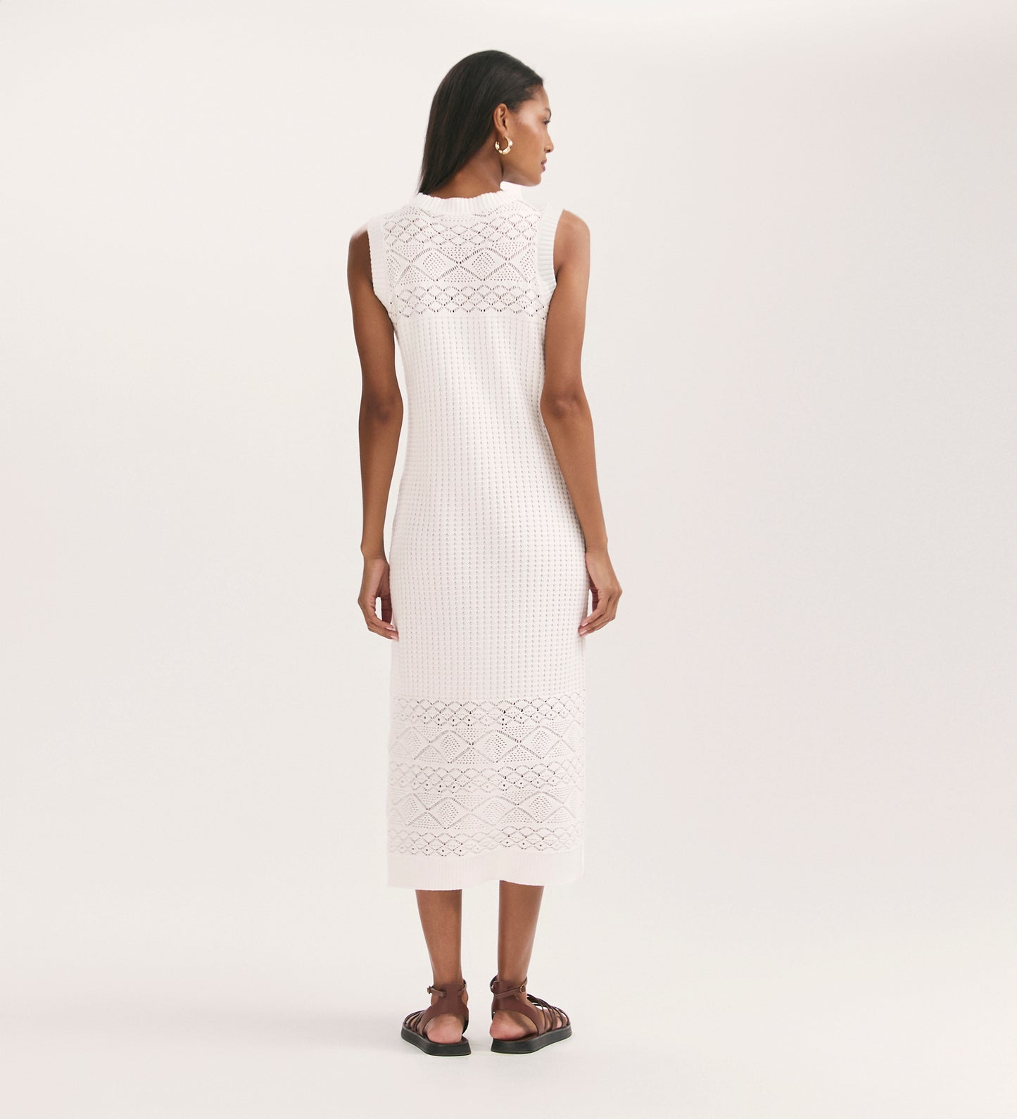 A woman stands with her back to the camera, wearing the Ravi Cream Sleeveless Knitted Midi Dress. The relaxed-fit, ankle-length dress features lace and crochet details. She accessorizes with hoop earrings and black sandals against a plain background.