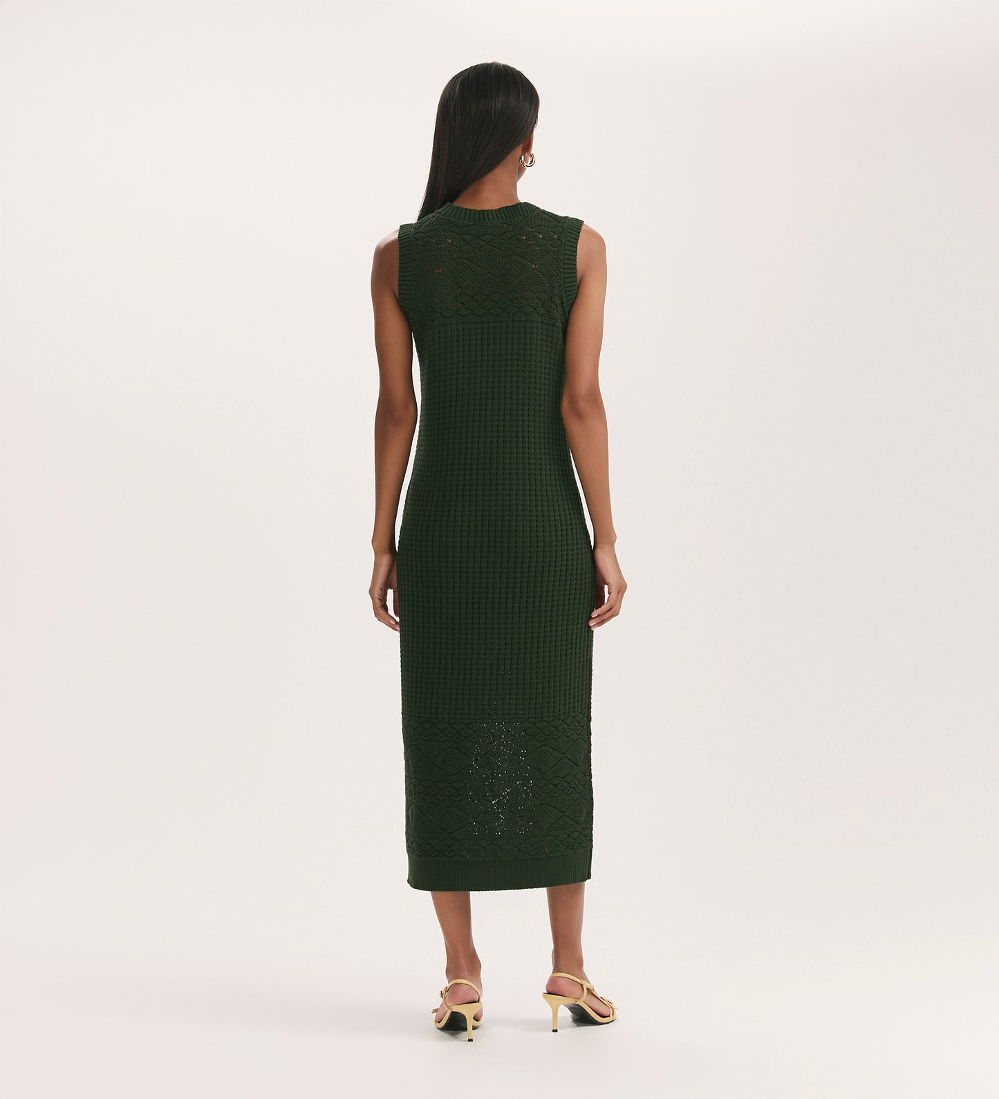 A woman stands with her back to the camera in the Ravi Khaki Sleeveless Knitted Midi Dress, a relaxed-fit midi style, paired with light-colored high-heeled sandals against a plain white background.