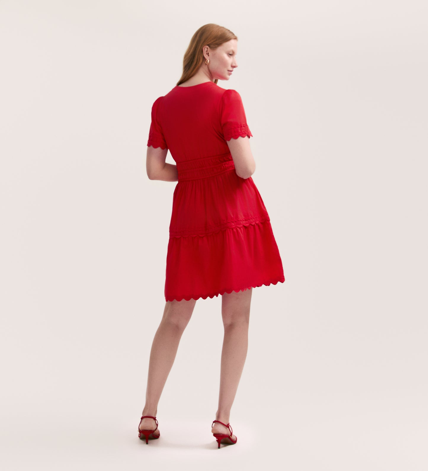 A woman faces away, wearing the Reece Red Short Dress—a short-sleeved, knee-length cotton dress with scalloped edges—paired with red heeled sandals against a plain, light background.