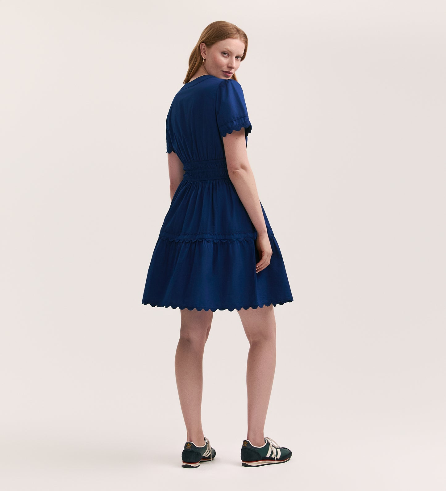 A woman with long, light brown hair models the Reece Indigo Ric Rac Trim Cotton Short Dress, a fit and flare style with a scalloped hem, paired with black sneakers, standing against a plain light background looking over her shoulder.